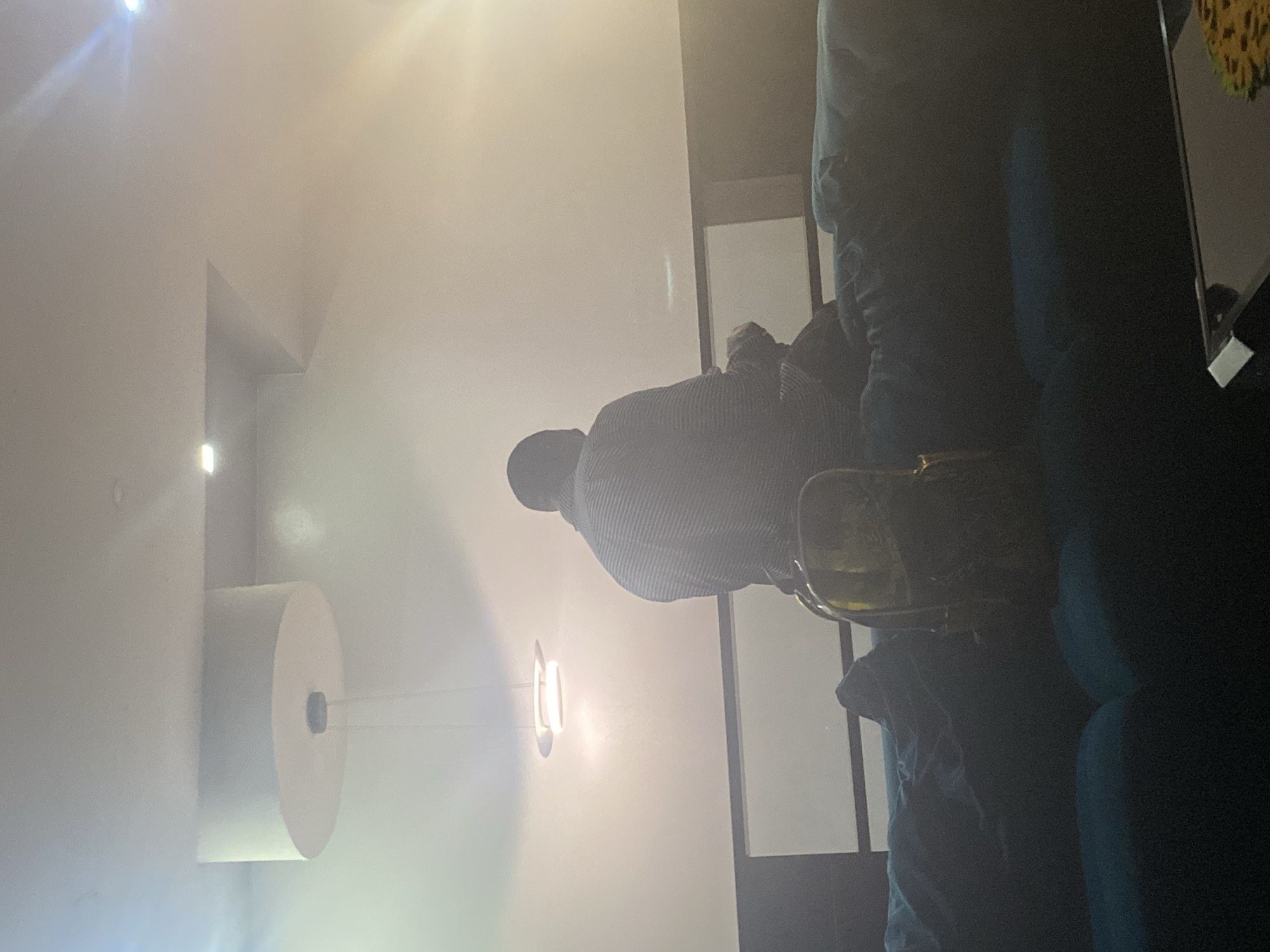 A man in a striped shirt is seated on a blue sofa. There is a lamp on the wall. The location is a room in an unknown city.