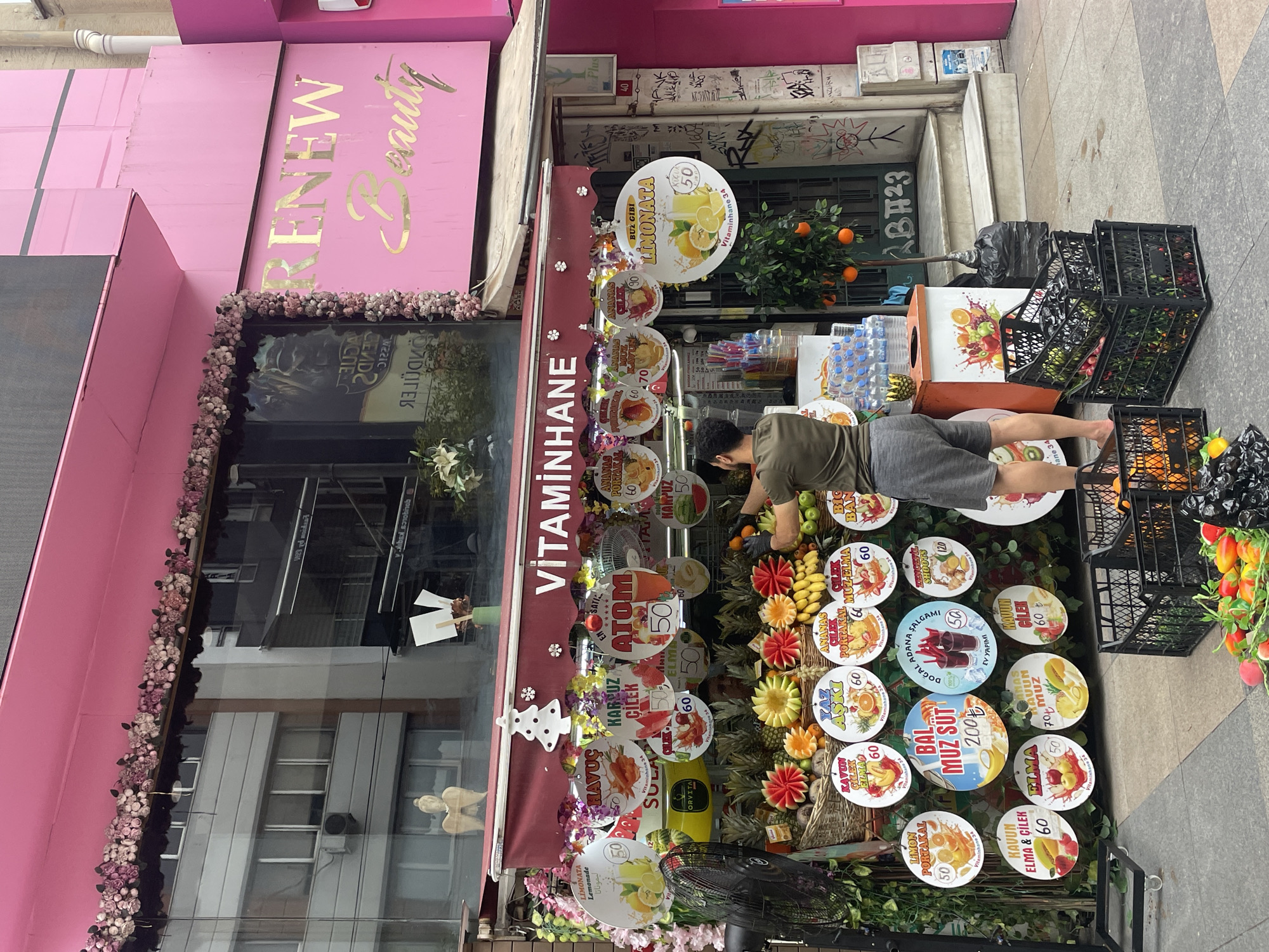 The image depicts a vibrant fruit juice stand in Kadıköy, Turkey, adorned with pink floral decorations and a bright pink awning. The stand is labeled "VİTAMİNHANE," indicating it serves vitamin-rich beverages. A man, wearing a green t-shirt and gray shorts, is actively preparing juices, his hands gloved as he handles oranges. He stands amidst an abundant display of fresh fruits, including pineapples, watermelons, bananas, and strawberries, artistically arranged. Colorful circular signs with prices and names of various juices, such as "Limonata," "Kavun Muz Süt," and "Ananas Portakal," are prominently displayed. Behind the fruit stand, stacks of black crates filled with produce and water bottles are visible. The background reveals a building with "RENEW Beauty" signage in gold lettering on a pink facade, suggesting a commercial area. The scene is bustling with activity, showcasing the lively atmosphere of a Turkish marketplace.