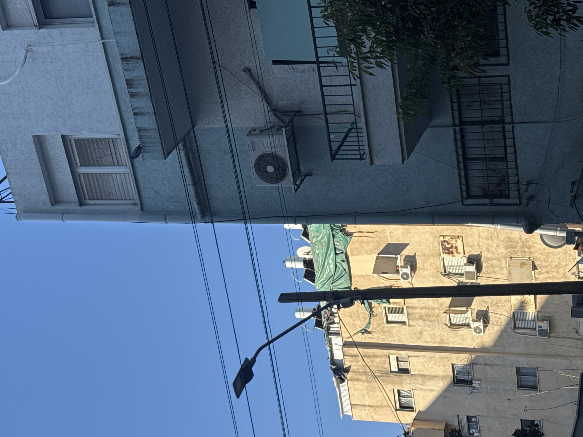 The image shows a street scene in Tel-Aviv, Israel, viewed from a tilted perspective. Two buildings are visible on opposite sides of the frame. The building on the top of the image has a pale blue facade, with windows, a fire escape ladder, and an air conditioning unit mounted on the exterior wall. The building on the bottom has a light-yellow facade, with windows and numerous air conditioning units. Power lines and a lamppost intersect the blue sky. A green tarp covers part of the bottom building. The overall impression is one of urban density.