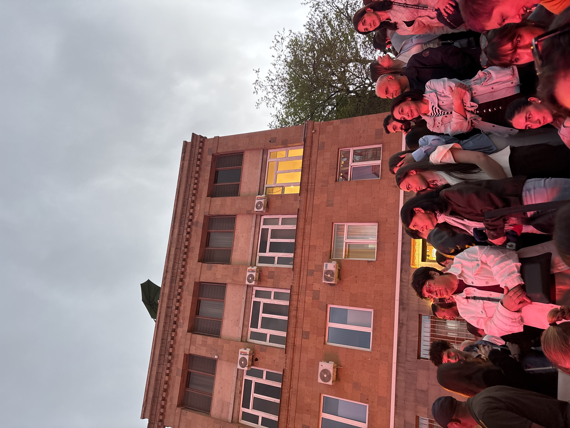 An outdoor scene captures a large crowd of people attending an event in Yerevan, Armenia, under an overcast sky. The image is taken from a low angle, looking up at a multi-story building and the faces of numerous individuals gathered on what appears to be a raised platform or balcony to the right.
The central subject is a tall, reddish-brown stone or brick building, possibly an apartment complex or office building, with numerous windows of varying styles and several white air conditioning units mounted on its facade. One window on an upper floor glows with a warm yellow light from within. A tree with sparse green leaves is visible behind the building's upper section, and a dark green umbrella is perched on the building's edge near the top left.
The crowd of people, mostly adults, fills the right side of the frame, their faces illuminated by a prominent red/orange light source coming from the right, suggesting evening or stage lighting. Many individuals are looking towards the left, indicating they are focused on something out of frame, possibly a stage or a focal point of the event. Their expressions are generally attentive. Some people wear jackets, sweaters, and shirts, with one individual's light-colored top partially displaying the text "MADE WITH LOVE." The overall atmosphere appears to be a public gathering or performance.