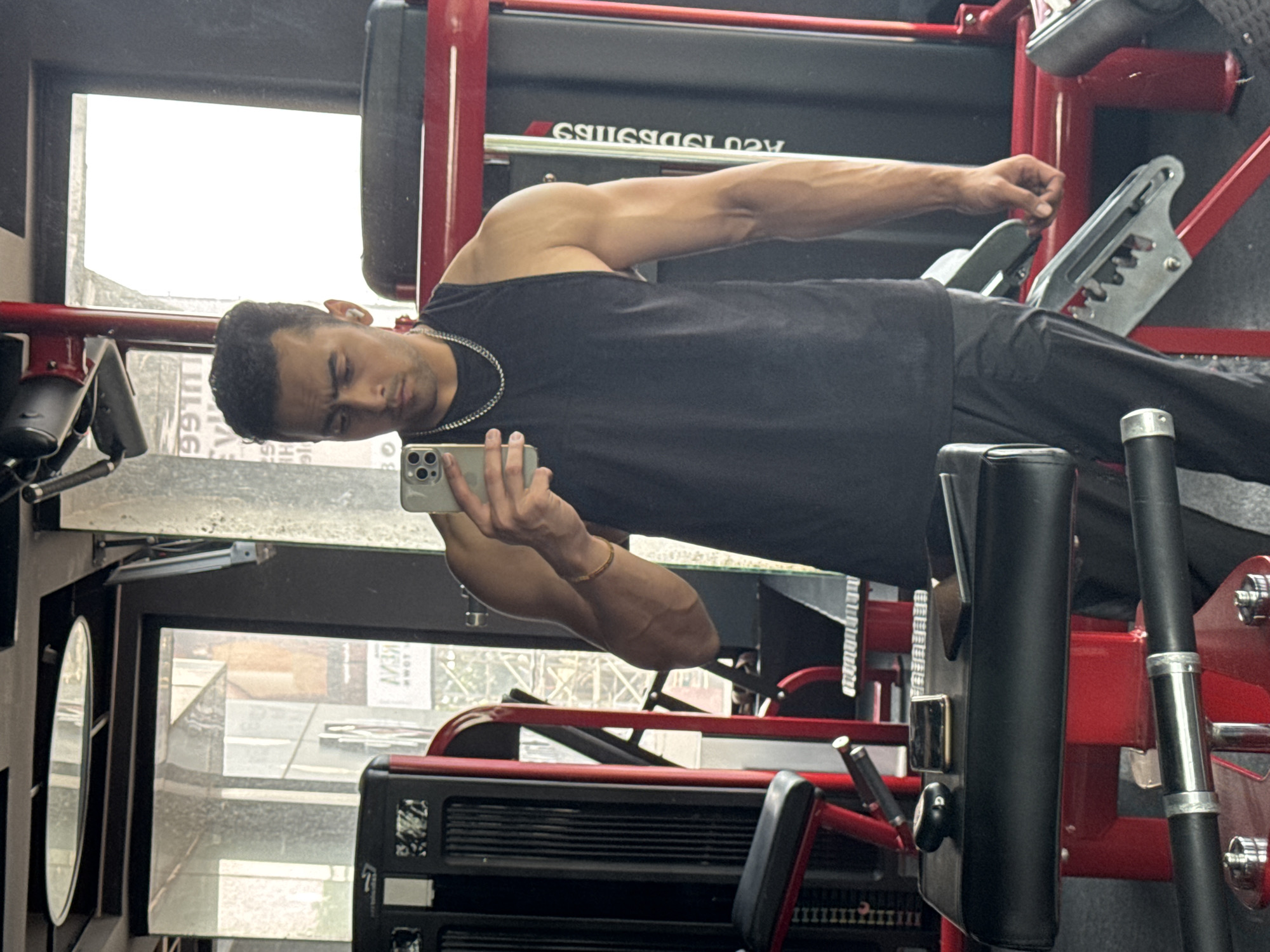 The image captures a muscular male subject taking a selfie with a light-colored smartphone in a gym setting. The man has dark hair, wears a black tank top, dark pants, a silver chain necklace, a golden bracelet on his right wrist, and a white earbud in his right ear. He holds the phone in his right hand, facing the mirror.
The environment is an indoor fitness center with prominent red and black exercise equipment. A weight stack machine is visible on the left, and another red-framed machine on the right. In the immediate foreground, a black padded bench or armrest holds a black wireless earbud case, a dark-colored smartphone, and a small, dark rectangular object. A metal bar is situated below the bench. The flooring is a dark, textured gym mat.
Behind the subject, a large window reflects the urban exterior, indicating daytime. The reflection reveals multi-story buildings, some with glass facades, and fragmented signage. Crucially, a reflective sign displays the word "TOWN" (visible as "NWOT" due to reflection) above which Bengali script is discernible. This linguistic identifier, combined with the urban backdrop, confirms the location as a gym within the city of Kolkata, India. Further fragmented text, such as "991T" and "EVI HLI", is also visible in the reflections.