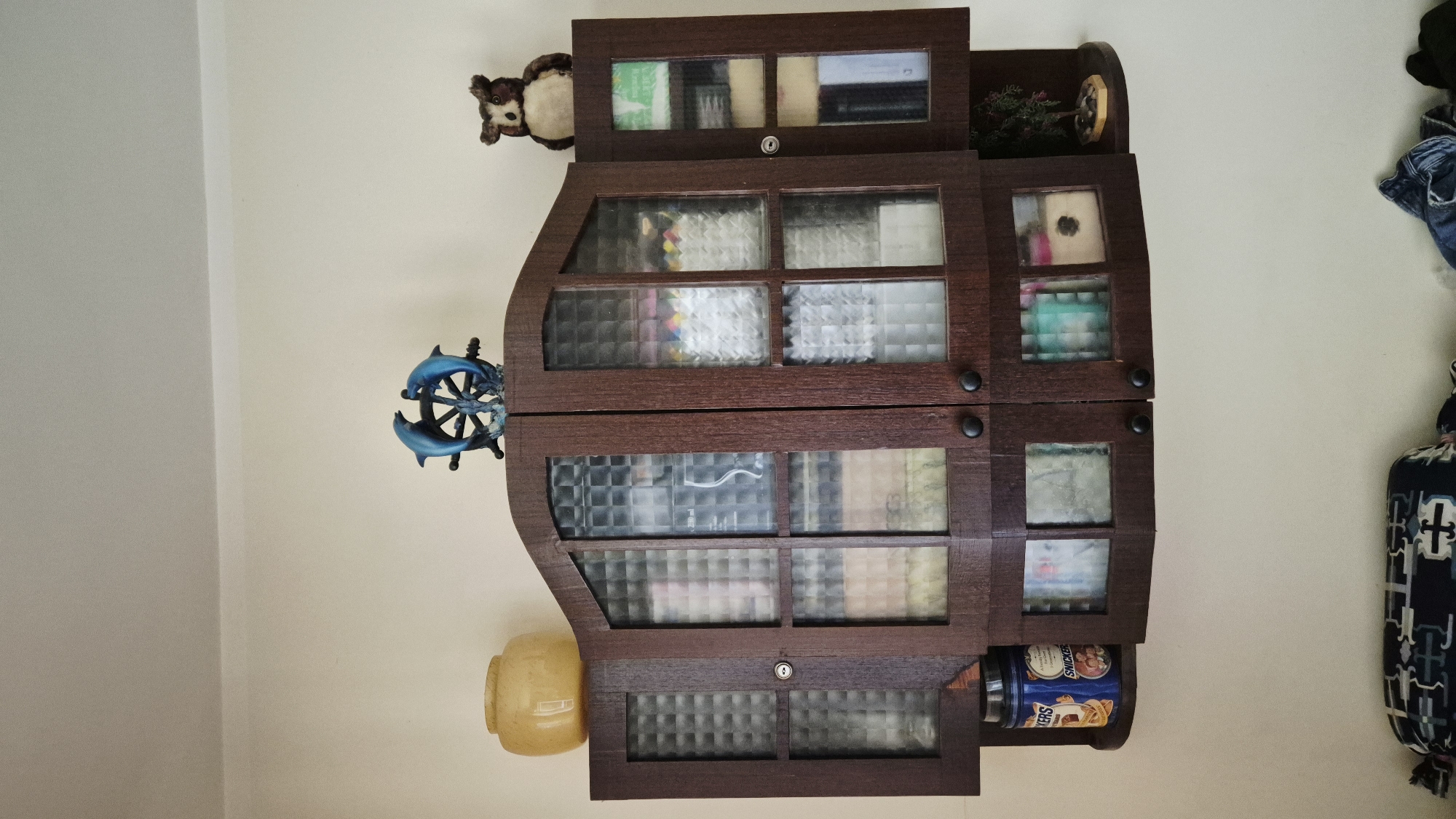 A wooden cabinet with glass doors is mounted on a light-colored wall. The cabinet is dark brown and has multiple compartments filled with various items. On top of the cabinet, there is a decorative sculpture of two blue dolphins on a base, and a plush owl toy. To the left of the cabinet, on a small shelf, sits a yellow glass jar. To the right, on another small shelf, is a miniature tree. In front of the cabinet, on the floor, are two folded items of clothing. The scene appears to be an indoor setting, possibly a room in Varena, Lithuania.