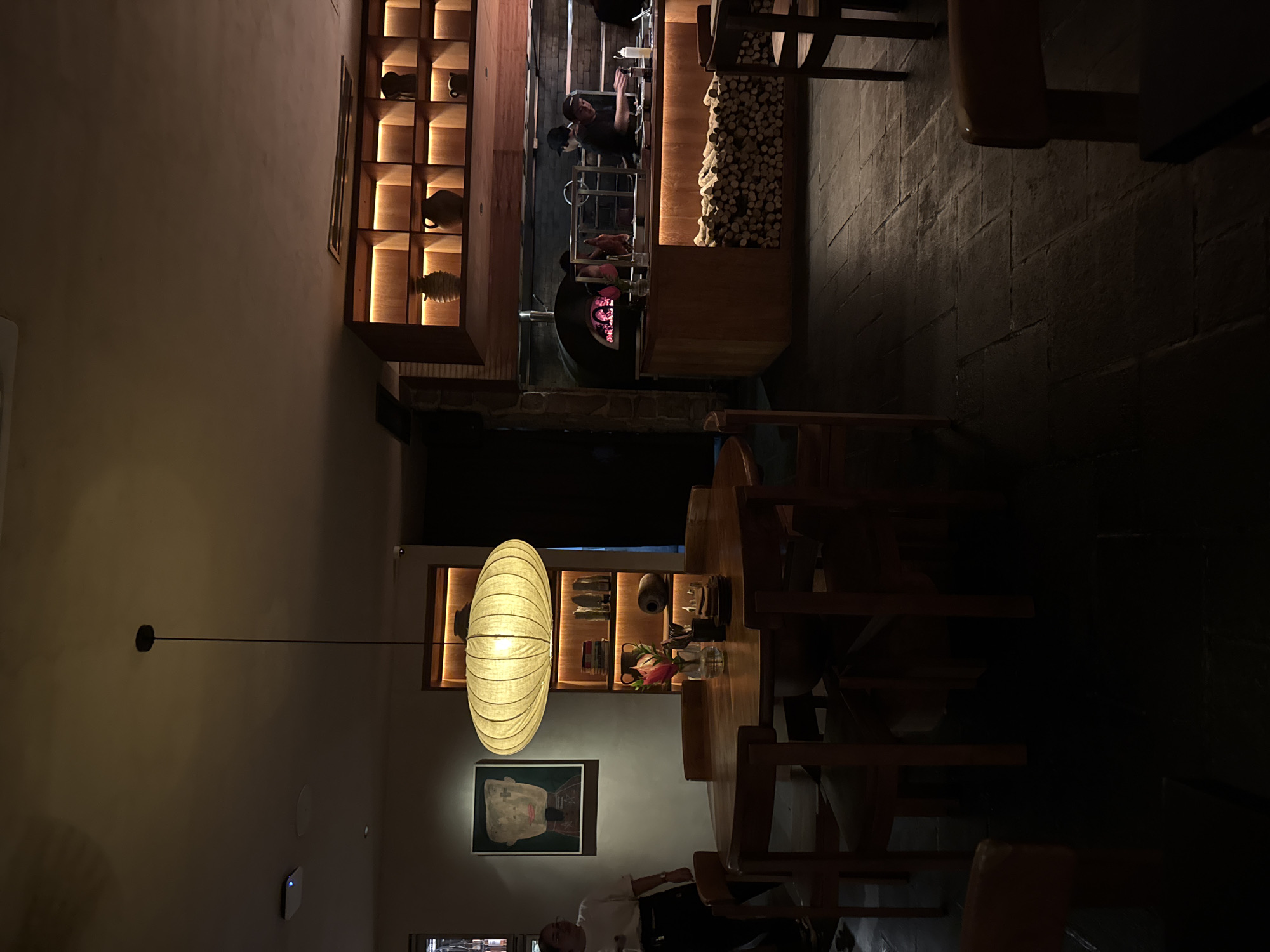 The image depicts the interior of a restaurant, likely located in North Kuta, Indonesia. The restaurant features a rustic, contemporary design with a visible wood-fired oven and open kitchen area staffed by at least two individuals. The dining area includes wooden tables and chairs, illuminated by an oblong pendant light fixture. The wall decor consists of shelving units displaying pottery and decorative objects, along with framed artwork. The lighting is warm and ambient, contributing to an intimate dining atmosphere.