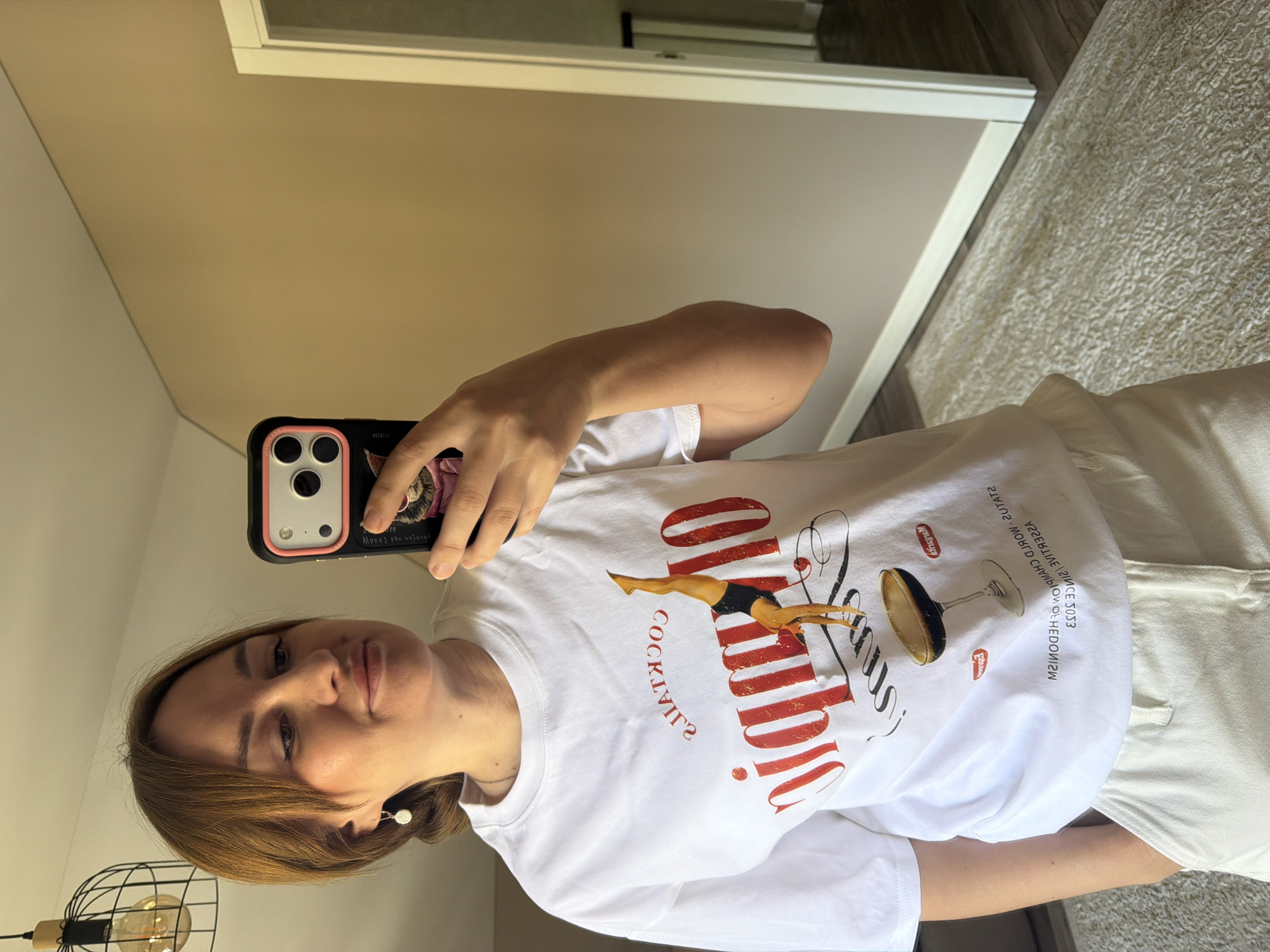 A woman is taking a selfie in a well-lit room, likely a bedroom, with light beige walls and dark wood flooring. She is wearing a white t-shirt with a graphic design featuring a diving figure and the text "Olympic Cocktails." She has her hair pulled back and is wearing a small silver earring. The room appears to be an indoor domestic setting, possibly in Malinovka, Russia, as there is a light fixture with a filament bulb hanging from the ceiling and a white doorframe is visible in the background, leading to another room. The overall ambiance is casual and personal.