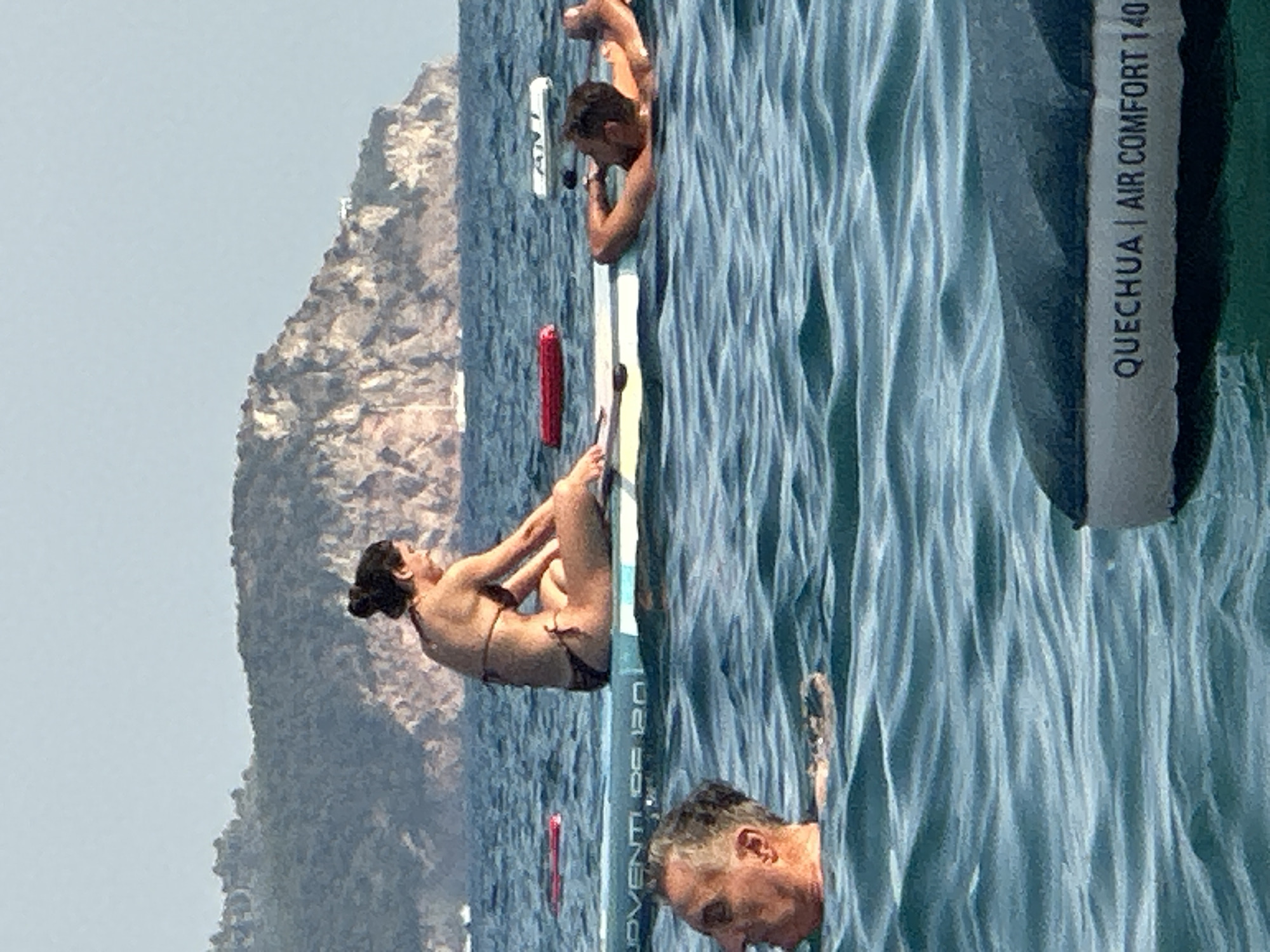 The image shows a sunny day at sea with a rugged, rocky island in the background. In the foreground, several people are in the water. A woman in a bikini is sitting on a paddleboard, and a man is partially submerged in the water next to her. Another man is floating on his back in the water, looking up at the sky. A paddleboard with the text "QUECHUA | AIR COMFORT 140" is visible at the bottom of the frame. Red and white buoys are also present in the water. The water is a vibrant blue, with small waves. This scene is likely taking place in the waters near the city of Boreti, Montenegro, given the presence of the distinctive rocky coastline.