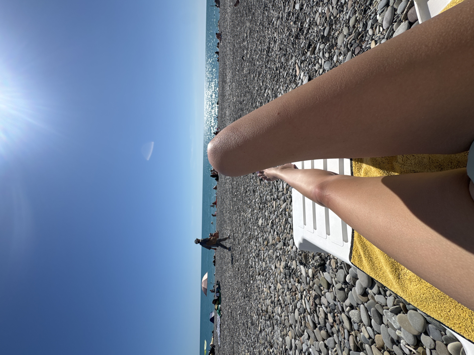 A first-person perspective shot shows a person relaxing on a white lounge chair on a pebble beach in Sochi, Russia. The camera is positioned to capture the lower half of their legs and feet, adorned with purple nail polish. A bright yellow towel is draped over the lounge chair. The beach is covered in grey pebbles, with the turquoise waters of the Black Sea visible in the background. Several other beachgoers are scattered across the shore, some under umbrellas. The sun is shining brightly, creating a shimmering effect on the water's surface and casting a shadow on the person's legs. The sky is a clear, vibrant blue, with the sun positioned high and to the right, its rays radiating outwards. The scene depicts a typical summer day at the beach.