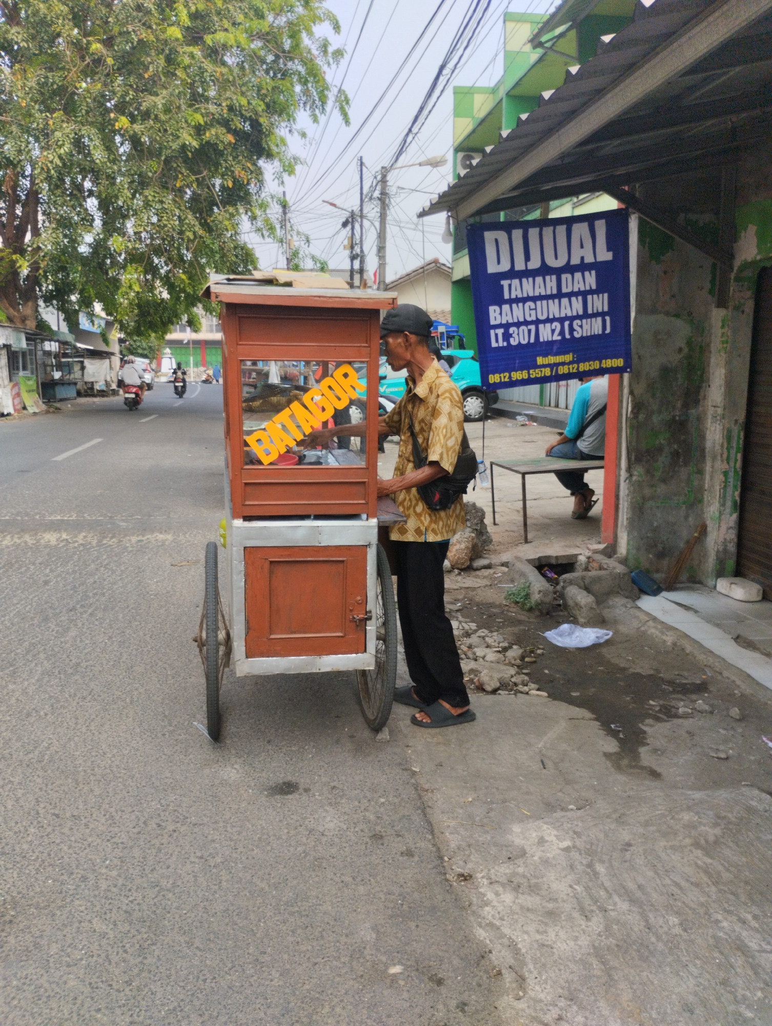 A man in a patterned shirt and black pants is standing next to a food cart on a street in Bekasi, Indonesia. The food cart has a wooden top and a metal base with two wheels. The word "BATAGOR" is written in large yellow letters on the side of the cart. Behind the man, a blue sign on a building reads "DIJUAL TANAH DAN BANGUNAN INI LT. 307 M2 (SHM)". In the background, there are other buildings, trees, and a few motorcycles on the street. The man appears to be preparing or serving food.