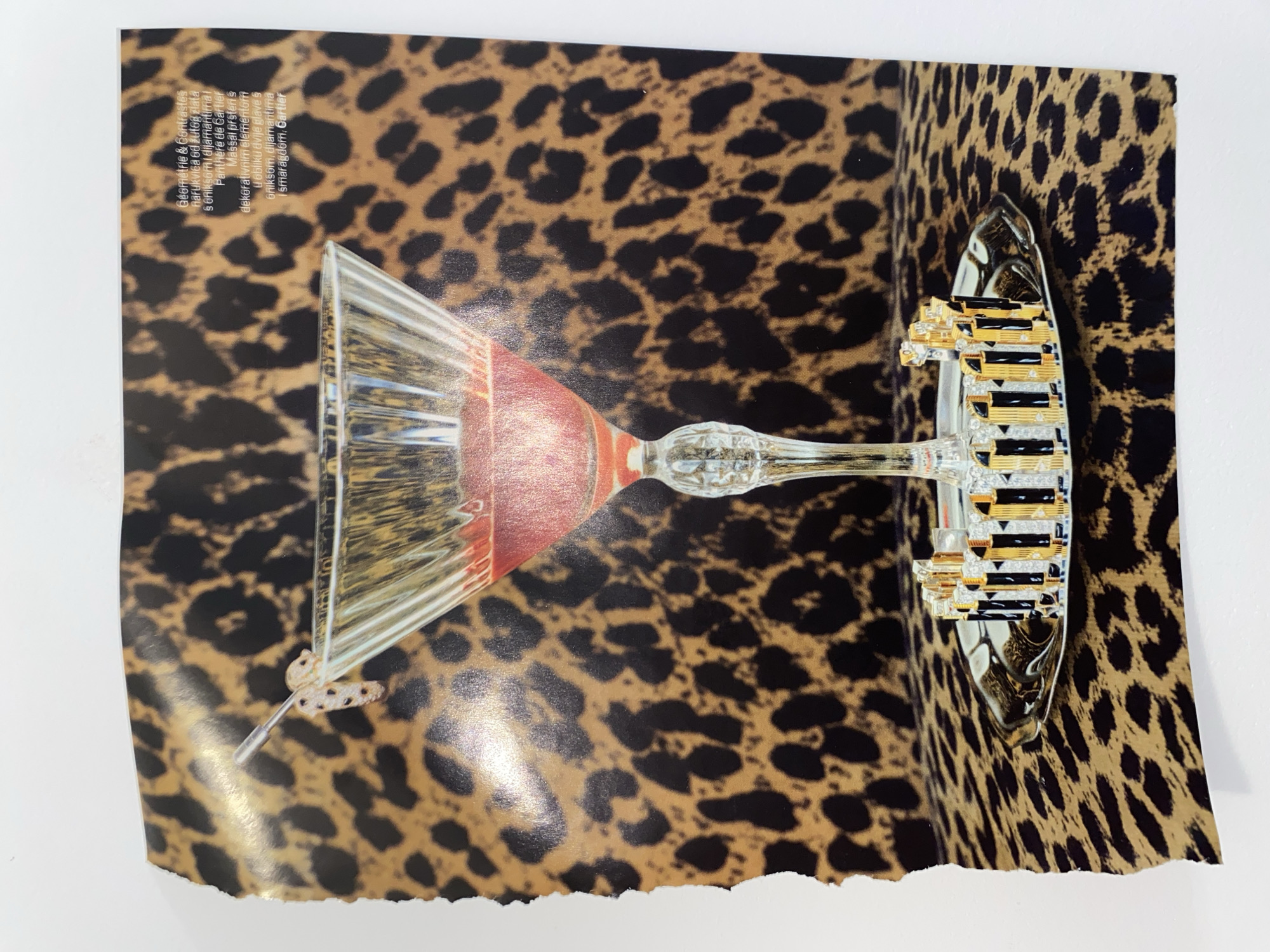 A photograph captures a page from a magazine or printout, featuring a leopard print background in shades of brown and black. The page is laid flat on a plain white surface, with its top and bottom edges slightly curled, and the bottom edge appearing roughly torn.
Prominently displayed on the page, against the leopard print, are two main objects. On the left, a clear stemmed glass, possibly a martini or cocktail glass, is depicted. Its bowl has a reddish-orange hue towards the bottom, and the stem is intricately detailed. On the right, a distinctive, multi-layered object is shown. It appears to be a decorative structure, possibly a piece of jewelry or an architectural miniature, crafted in gold and black with sparkling clear accents, resembling stacked rectangular elements.
In the upper left corner of the page, text is visible, oriented sideways. The text, largely in Croatian, reads: "Géométrie & Contrastes narukvica od žutog zlata § oniksom i dijamantimai Panthere de Cartier Massal prsten s dekorativnim elementom u obliku dvije glave s @niksom, dijamantima I smaragdom, Cartier." This translates to "Geometry & Contrasts, yellow gold bracelet with onyx and diamonds, Panthere de Cartier Massal ring with decorative elements in the shape of two heads with onyx, diamonds, and emeralds, Cartier." This suggests the image is an advertisement or editorial for Cartier jewelry.