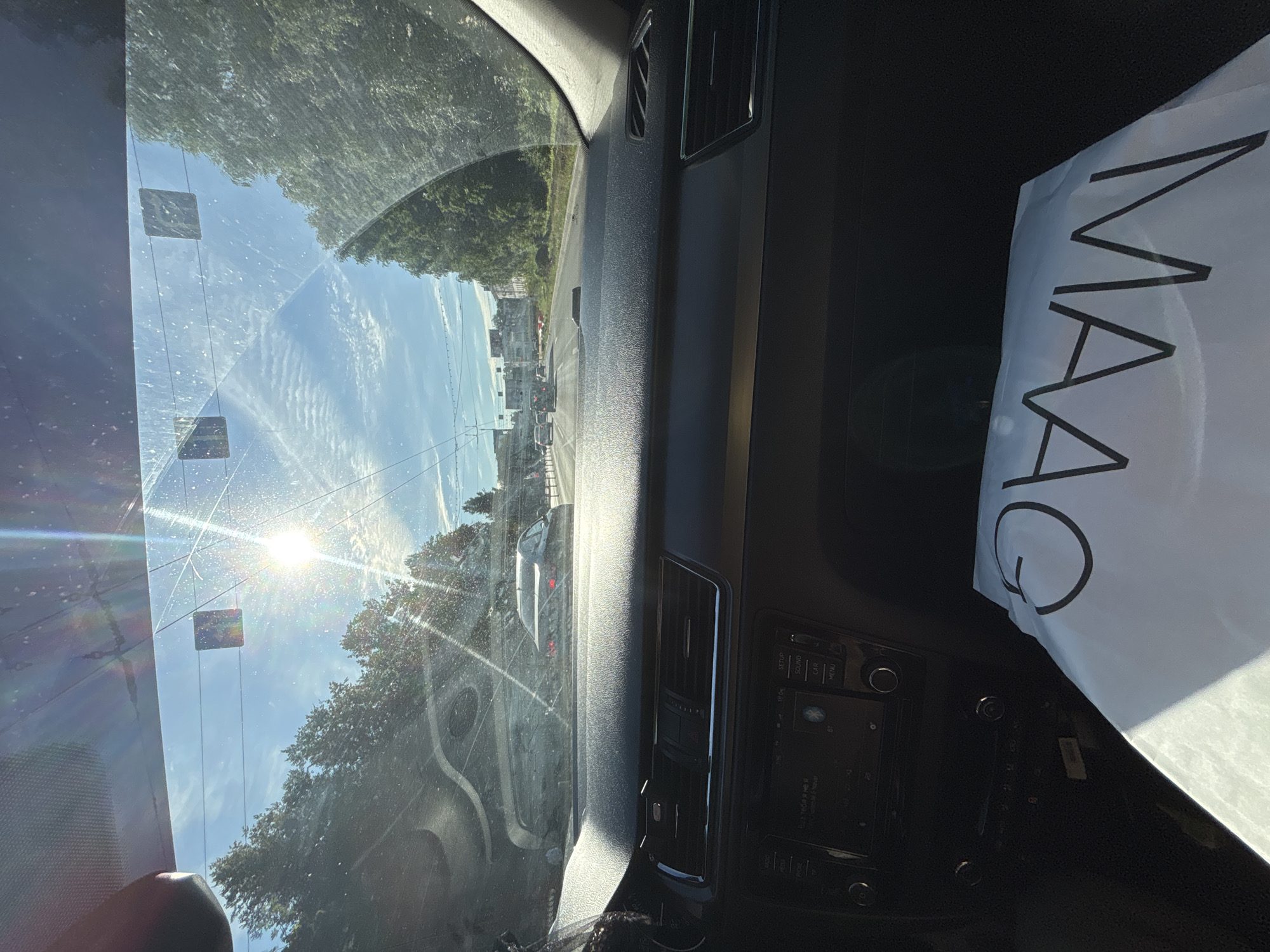 The image shows the interior of a car, looking forward through the windshield. The sun is bright and glares through the windshield, creating lens flare. Trees line the sides of a road, and several cars are visible in front. In the foreground, on the dashboard, is a white bag with the word "MAAG" printed on it in black lettering. The scene appears to be a daytime shot, likely taken from the passenger seat or the driver's seat while the car is in motion on a road in Rusanovo, Russia.