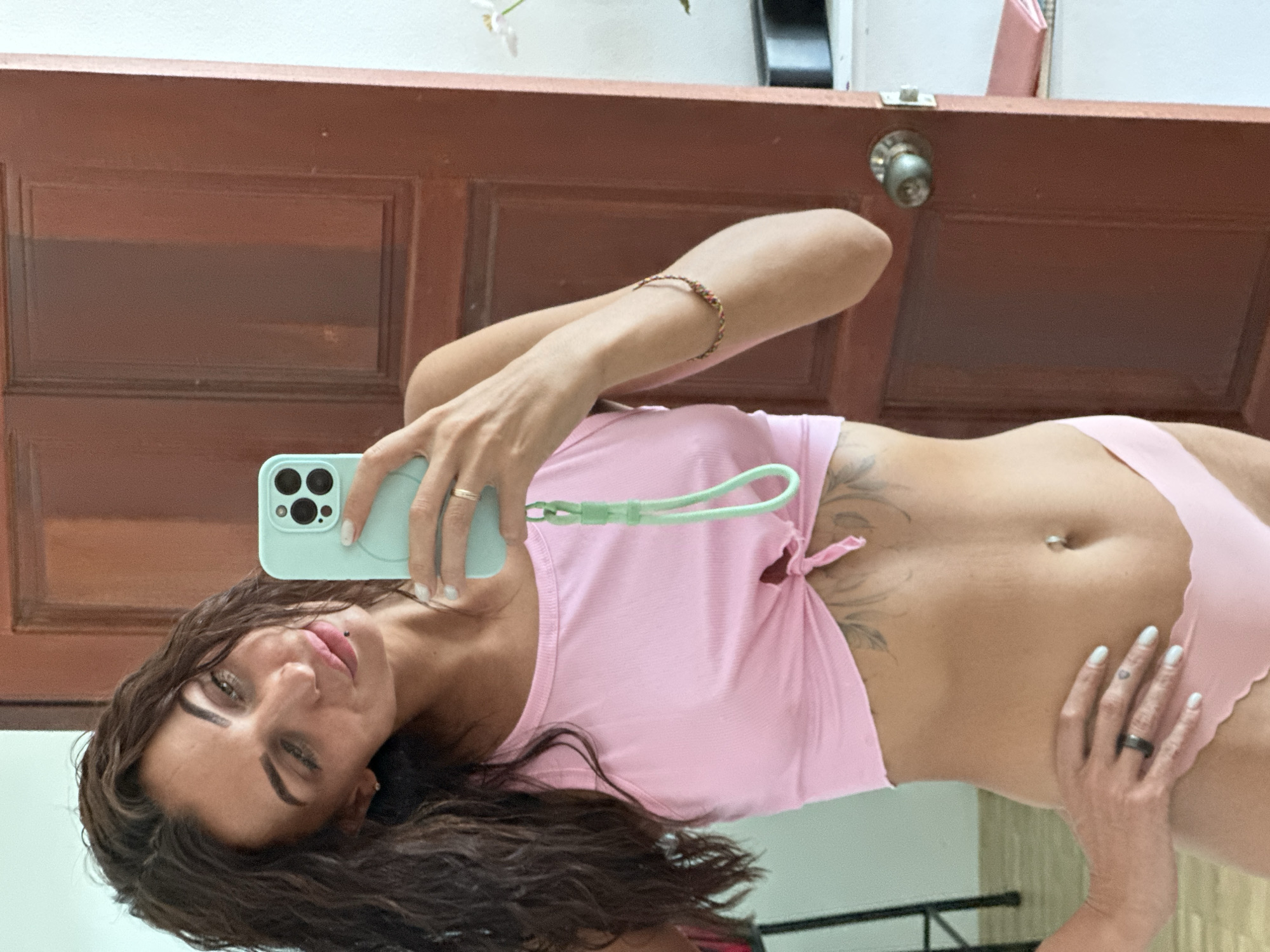 The image shows a woman taking a mirror selfie indoors, possibly in Ko Samui District, Thailand, given the context. She is wearing a pink crop top tied at the front, exposing her midriff, and pink bottoms. A tattoo is visible on her stomach, and she has a navel piercing. She is wearing jewelry on her left hand, including a dark ring, and a thin bracelet on her right arm. Her hair is dark and wavy, and she is looking at her reflection in a mirror. The background includes a wooden door with a doorknob and a white wall. A phone in a light blue case is being held by the woman to take the selfie.
