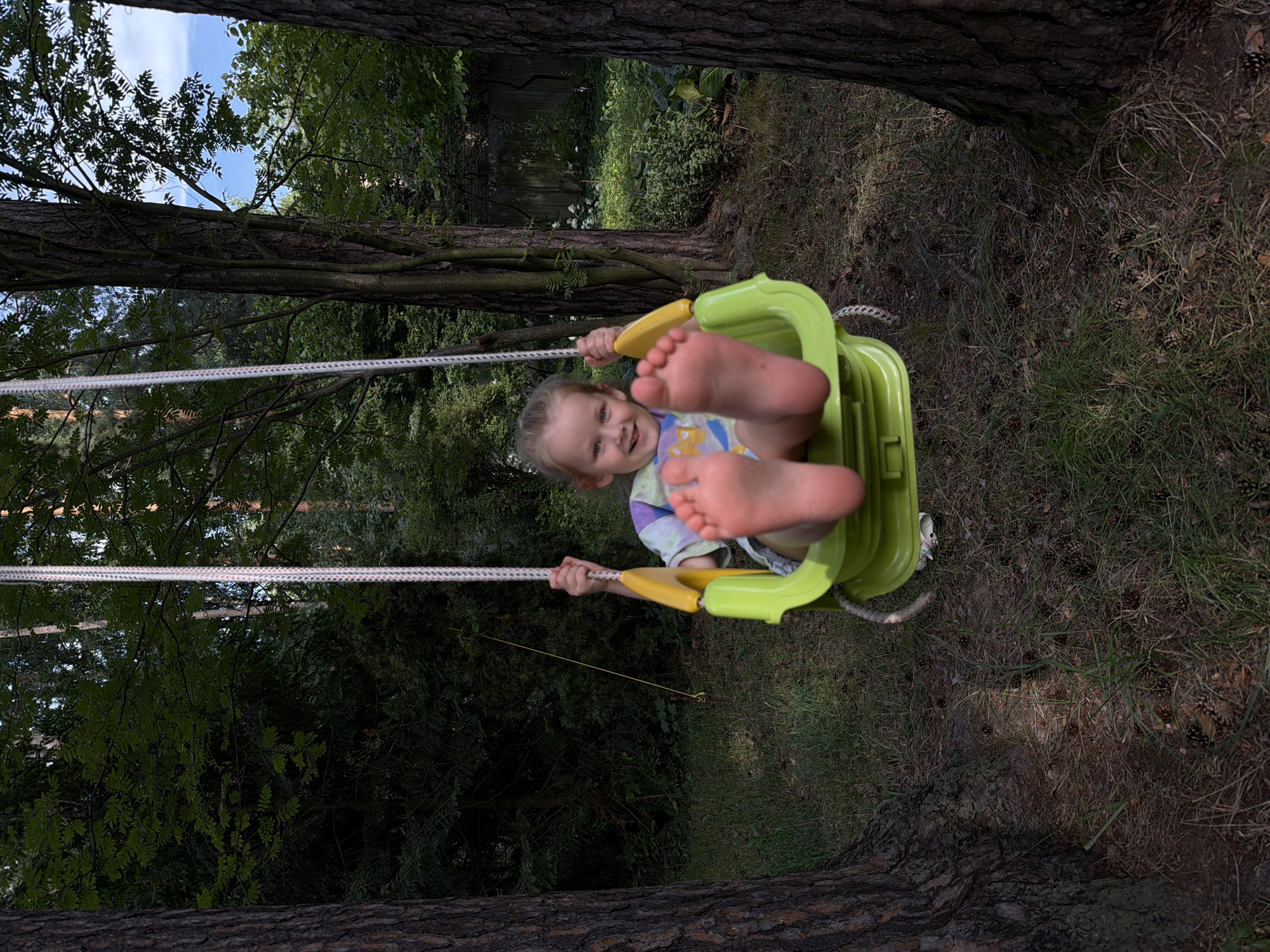 A young child is swinging outdoors, viewed from a low angle. The child, with fair skin and blonde hair, is sitting in a lime green swing with a yellow bar. They are wearing a colorful t-shirt with a tie-dye pattern. The child is smiling and looking towards the camera, with their feet prominently displayed. The swing is suspended by thick ropes attached to a tree. The surrounding environment is a wooded area, with tall trees and dense foliage. The ground is covered in grass, pine needles, and scattered pinecones. Sunlight filters through the leaves, suggesting it is daytime. The overall mood is playful and joyful. The location is identified as Udelnaya, Russia.