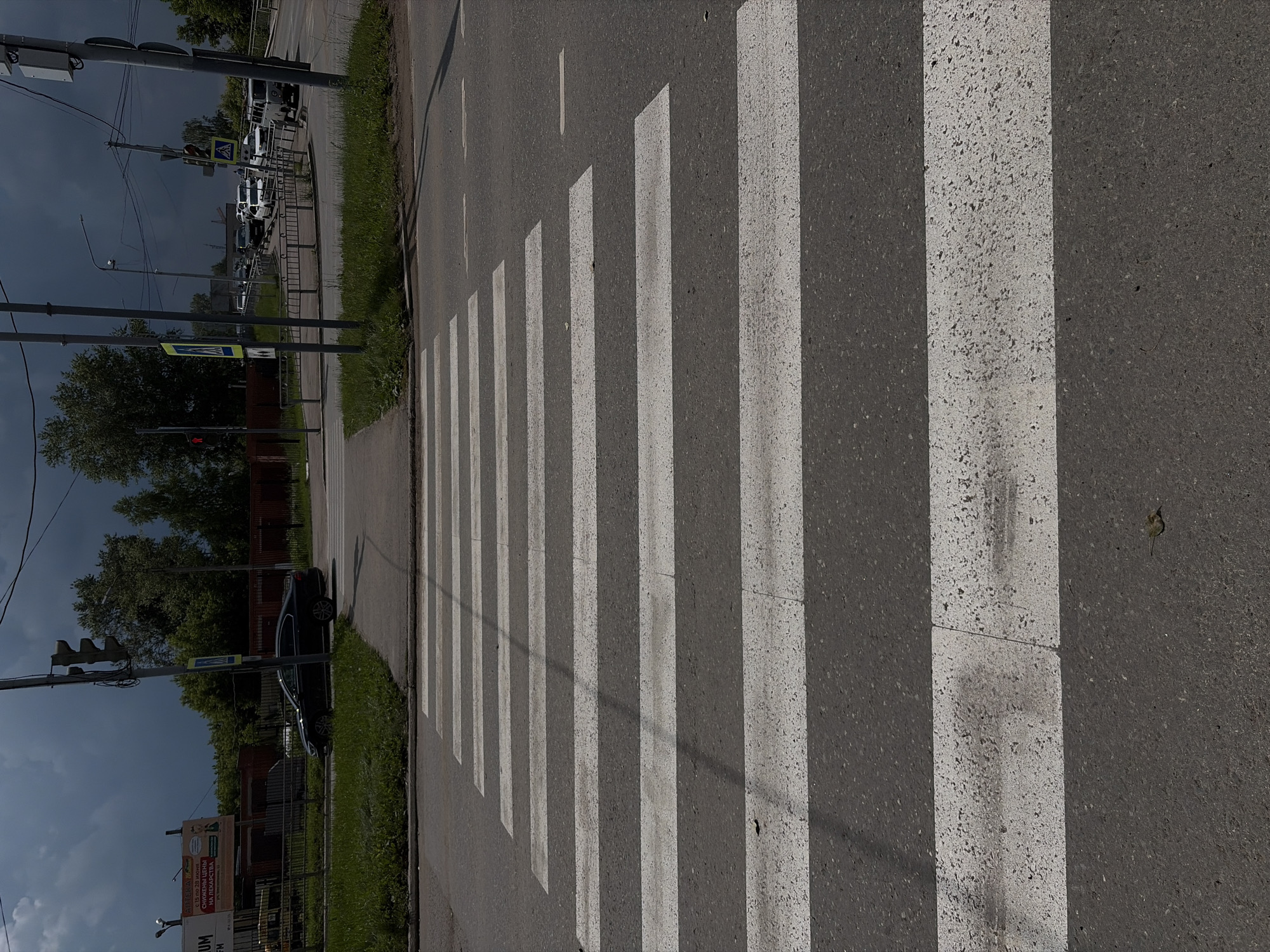 The image depicts a crosswalk on an asphalt road, with multiple white stripes indicating the pedestrian crossing. In the background, there are trees, a fence, and some buildings. A dark, cloudy sky looms overhead. A black car is parked on the left side of the road, facing away from the viewer. Traffic lights and pedestrian crossing signs are visible near the intersection. There is also some traffic in the distance, including a white van and other cars. The scene appears to be an urban or suburban environment.