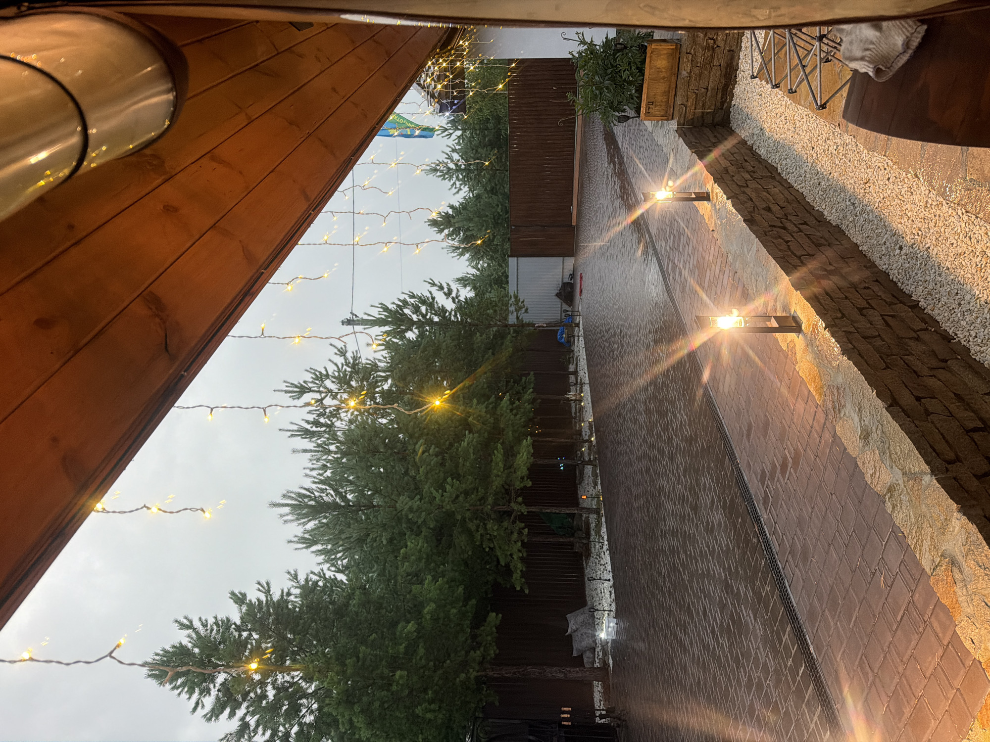 The image depicts an outdoor paved area, likely a courtyard, with a wooden overhang extending into the frame from the upper right. The ground is paved with reddish-brown interlocking bricks and appears wet, suggesting recent rain. A metal drain grate runs across the paving. Along the edge of the paved area, a low wall constructed of stone and brick is visible. This wall is illuminated by two modern, rectangular, solar-powered pathway lights, each emitting a warm yellow glow. Beyond the wall, a narrow strip of white gravel is present.
A line of trees, predominantly pine trees, with dense green foliage, forms a backdrop to the scene, extending across the width of the image. Behind the trees, a dark wooden fence encloses the property. In the distance, a lighter colored metal fence is visible. The sky is overcast and a pale grey, indicating a cloudy day or dusk.
Decorative strings of small, warm white fairy lights are draped from the wooden overhang, with some strands extending towards the trees. A flagpole with what appears to be the Ukrainian flag is visible on the right side of the image, attached to a building structure. A planter box containing green foliage sits on top of the stone and brick wall on the right. In the foreground, to the far right, a dark wooden surface, possibly a table or bench, is partially visible, with a folded grey blanket resting on it.
There are no people present in the image. The overall impression is one of a well-maintained outdoor space, possibly a residential property in a rural or suburban area, with decorative lighting suggesting an evening or early morning setting. The location is identified as Kolyutkino, Russia.