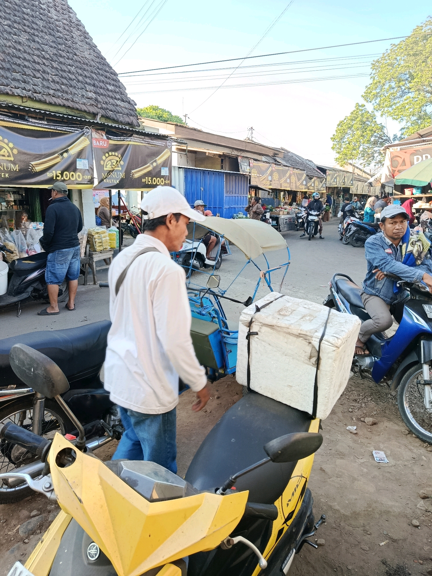 The image depicts an outdoor market scene in Bululawang, Indonesia. In the foreground, a yellow scooter is parked. Behind it, a person wearing a white long-sleeved shirt and blue jeans is walking away from the camera, carrying a strap over their shoulder. Further back, a man in a denim jacket sits on a blue motorcycle, looking towards the camera, with a patterned cloth draped over his arm. A large white cooler box is strapped to the back of a blue motorcycle. In the background, a row of market stalls with awnings line a dirt path. Several people are visible browsing or working in the stalls. The sky is bright and appears to be late afternoon. The overall atmosphere is that of a bustling local market.