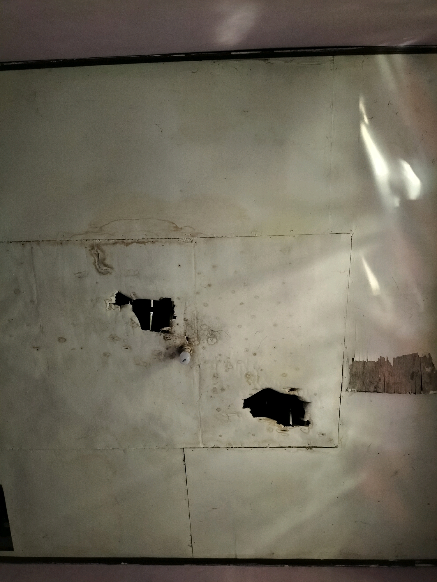 The image shows a damaged ceiling in a room. The ceiling is made of light-colored panels, some of which are discolored with water stains. There are two large holes in the ceiling. One hole is irregular in shape and located in the center of the image, revealing darkness within. The other hole, to the right of the first, is also irregular and exposes wooden material. Light streaks are visible on the ceiling, suggesting sunlight or artificial light reflecting off the surface. A white circular object, possibly a light fixture, is partially visible near the larger hole. The edges of the room, where the ceiling meets the walls, are visible at the top and bottom of the image, showing dark lines and a hint of purple or pink color on the walls. The scene is forensic in nature, documenting damage and deterioration within an indoor environment, potentially indicating a structural issue or a past incident. There are no people or discernible activities depicted. The image is taken from a low-angle perspective, looking upwards. Without further context, the specific location in Sambirejo, Indonesia, cannot be determined from the visual information alone.