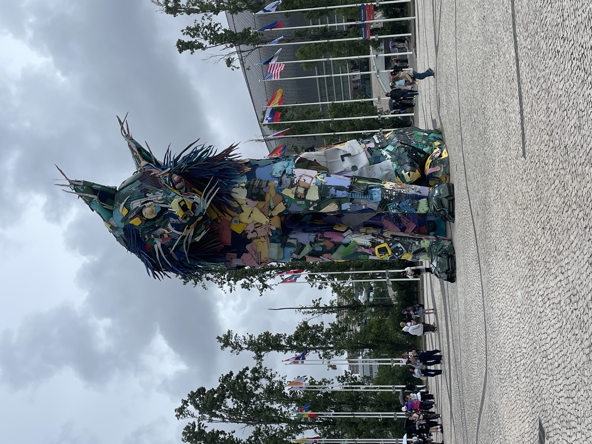 The image captures a large, colorful sculpture of a cat, constructed from various recycled materials, standing in a public square in Lisbon, Portugal. The sculpture is positioned prominently, with a building adorned with international flags in the background. The square is paved with a light-colored, patterned stone. Several people are visible, standing or walking near the sculpture and along the edges of the square. The sky is overcast with cloudy conditions. Trees line the square, situated between the flags and the building.