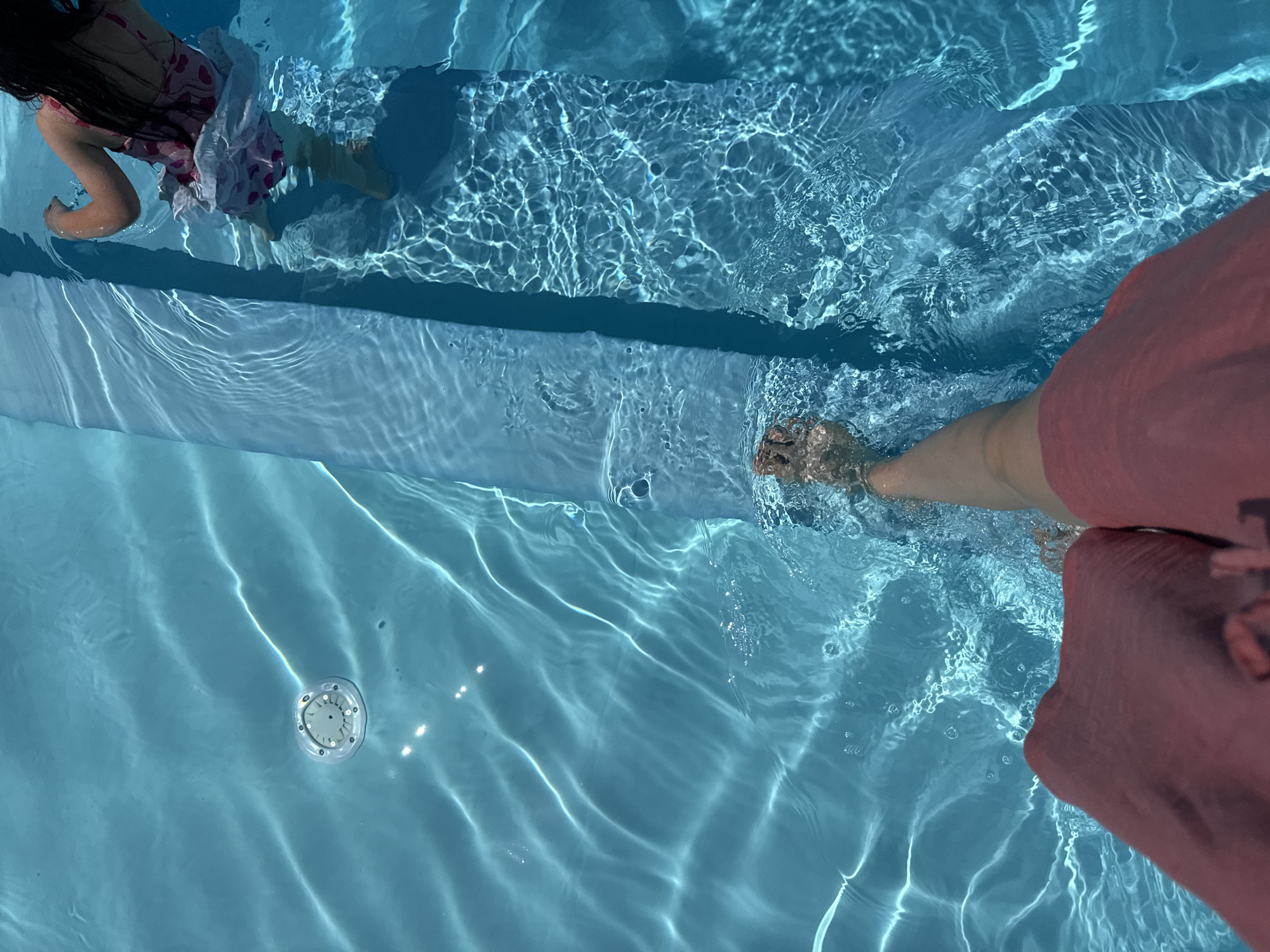 An overhead shot shows a person's feet entering a bright blue swimming pool. The person is wearing a pink garment and has their left leg submerged in the water. To the right, a child with dark hair is visible in the water, wearing a pink and white swimsuit. The child's arm is raised. The pool has a light blue edge and a circular jet is visible in the upper right quadrant. The water is clear and reflects sunlight, creating a rippling effect on the surface.