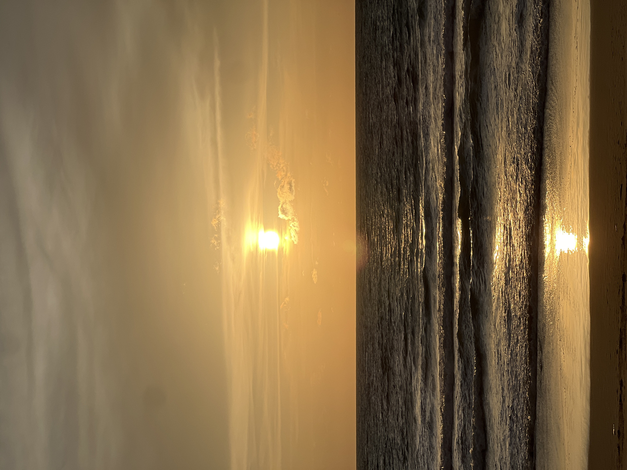 A vertical, eye-level shot captures a tranquil beach scene at sunset in Choeng Thale, Thailand. The sun, a bright orb with a halo of light, is positioned slightly left of center in the upper half of the frame, casting a warm, golden glow across the sky and sea. Wispy clouds, tinged with orange and yellow, are scattered across the horizon. The ocean, a deep, dark blue, is calm with gentle waves rolling towards the shore. Sunlight reflects off the water's surface, creating shimmering streaks of light. The wet sand at the bottom of the frame is a muted brown, reflecting the colors of the sunset. The scene is devoid of people, structures, or any man-made objects, emphasizing the natural beauty of the environment.