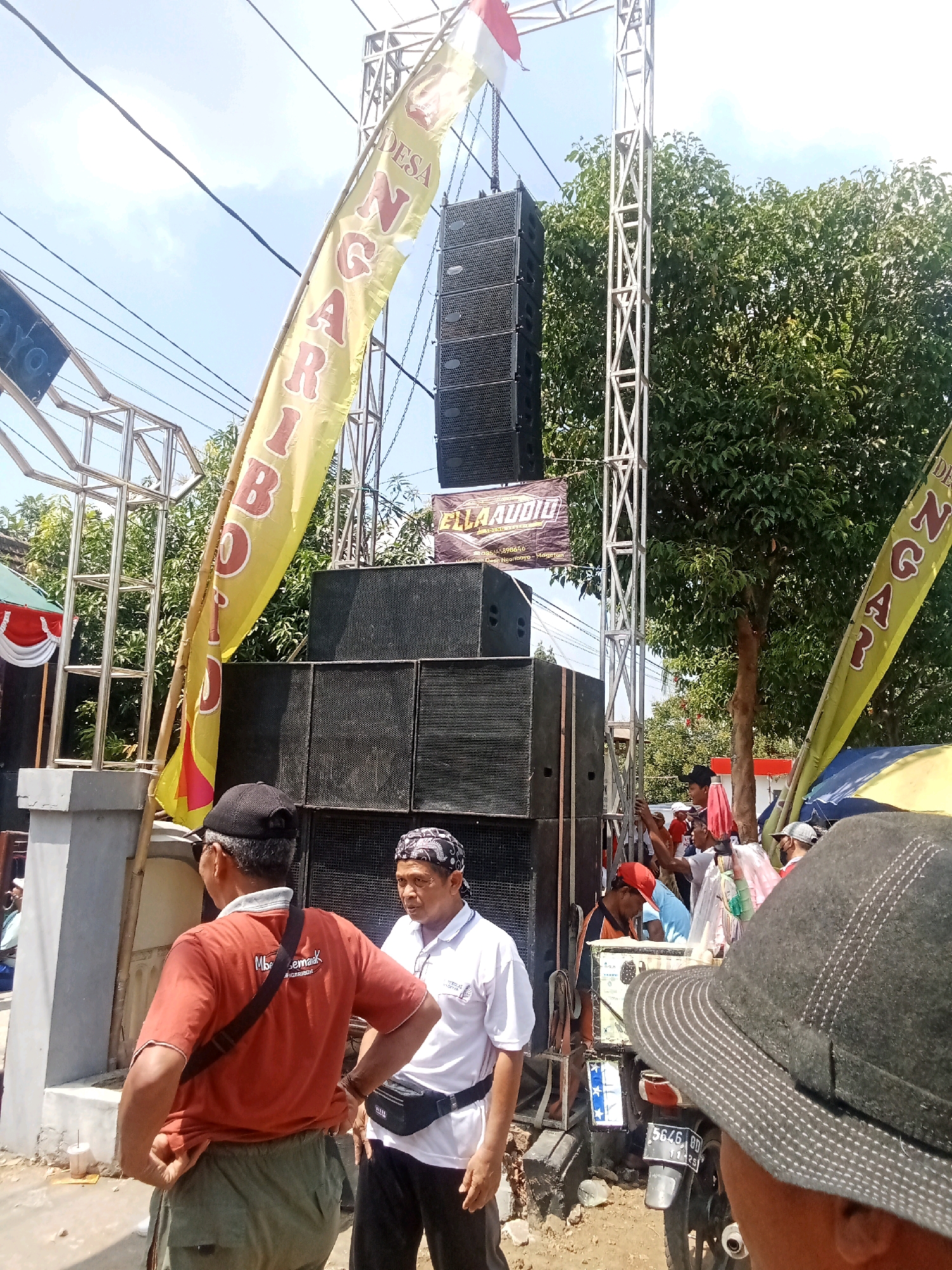The image depicts an outdoor event, likely a cultural or musical gathering, in the city of Ngariboyo, Indonesia. In the foreground, two men are visible. One man, wearing an orange t-shirt and green pants, is facing away from the camera, with his hand on his hip. The other man, in a white polo shirt and black pants, is facing the first man and looking towards him. A black fanny pack is worn around his waist.
Behind the men, a large sound system is set up, with several speaker cabinets stacked on top of each other and mounted on a tall metal truss structure. A yellow banner with red lettering is draped over the truss, displaying text that appears to be Indonesian. Another banner with "ELLA AUDIO" printed on it is also visible near the speakers. A few other individuals are seen in the background, some appearing to be part of the event crew or attendees.
The environment suggests a warm, sunny day, with a clear blue sky and lush green trees in the background. The ground appears to be dirt or gravel, indicative of an outdoor, possibly rural or semi-urban setting in Ngariboyo. Electrical wires are strung overhead, common in Indonesian urban and semi-urban areas. A motorcycle with a license plate is parked near some of the equipment. The overall scene suggests a lively event in progress or being set up.