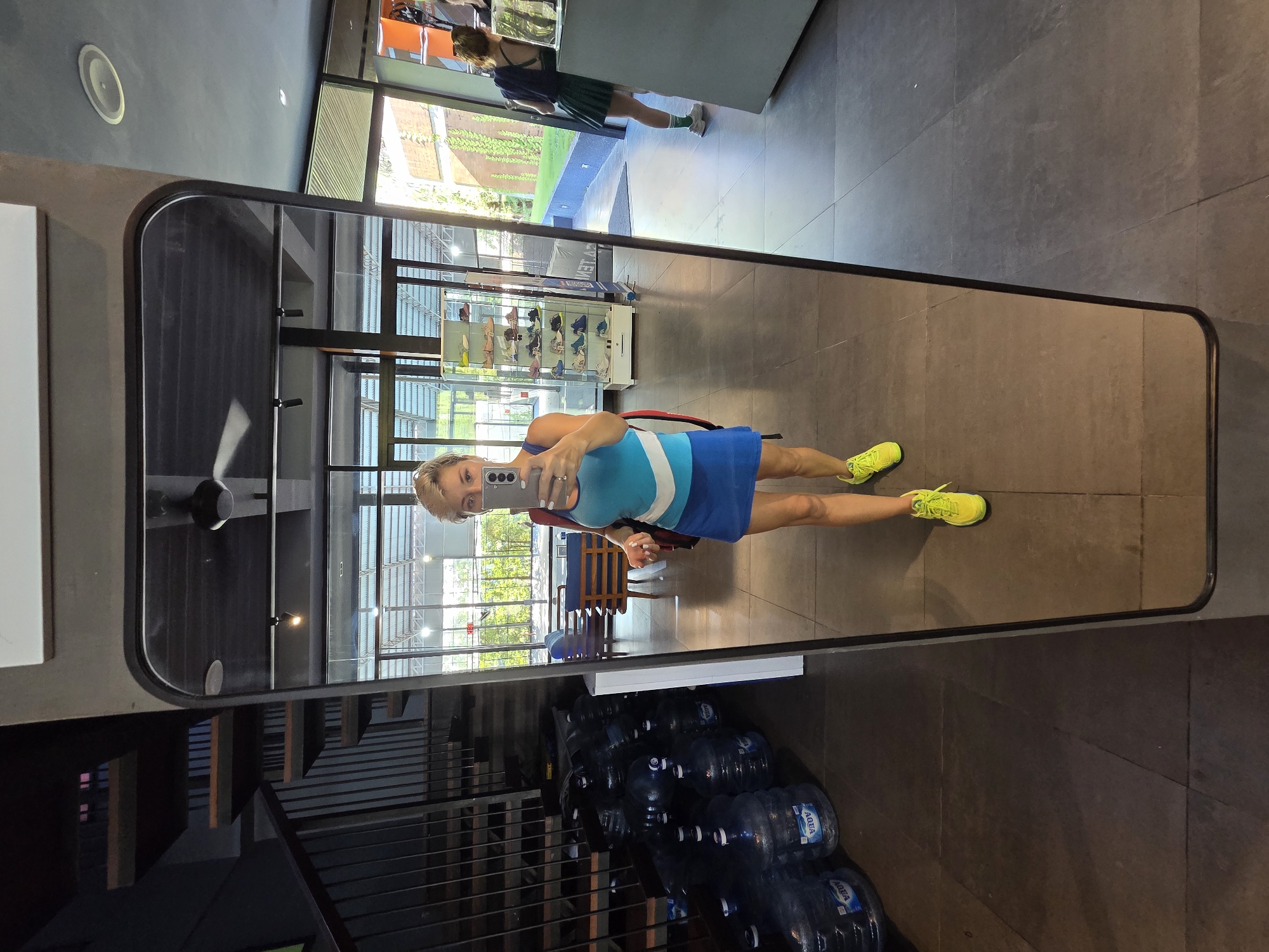 The image shows a woman taking a mirror selfie in what appears to be a store or gym. She is wearing a blue and white striped athletic dress and bright yellow sneakers. Her hair is blonde and styled in a short cut. She is holding a smartphone in her right hand, with her left hand resting on her hip.
The setting includes a large mirror that dominates the frame, reflecting the woman, the interior of the space, and some of the exterior through large windows. Inside, there are shelves with what seem to be shoes on display, a ceiling fan, and a staircase. Outside the window, greenery is visible, suggesting it is daytime. In the foreground, below the mirror, are several large water cooler bottles.
The woman's expression is neutral, and the overall mood of the image is casual. The lighting suggests it is daytime. The image was taken in North Kuta, Indonesia. Text is visible on the water bottles, reading "AQUA".