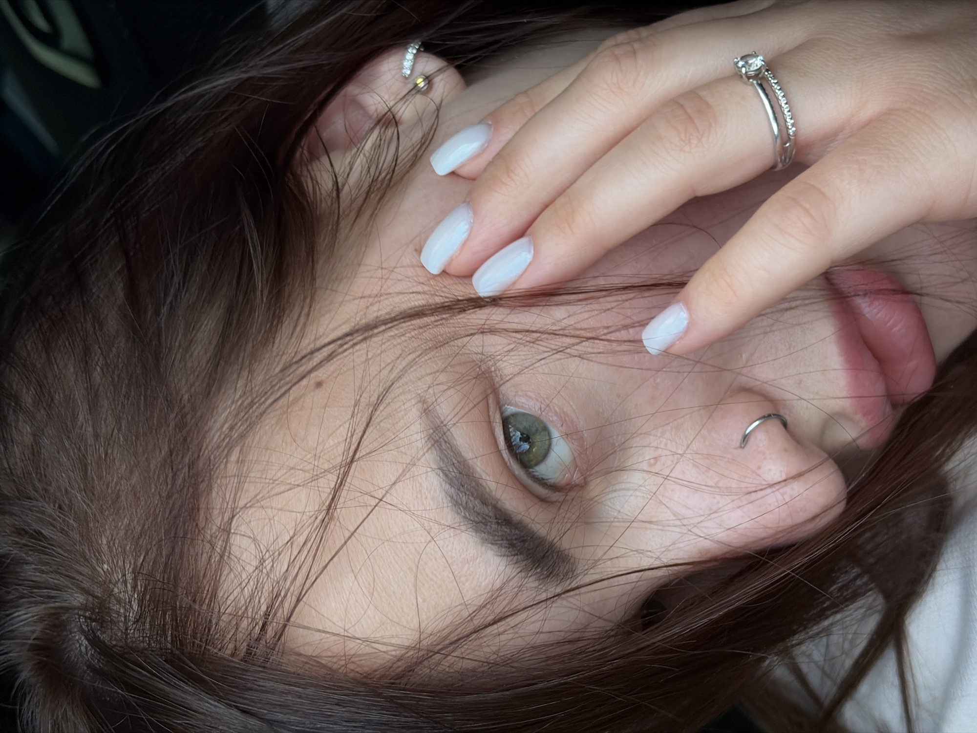 A close-up image shows a woman's face, partially obscured by her hand and hair. She has light skin, brown hair, and green eyes. She wears a ring on her finger, a nose ring in her left nostril, and an earring in her left ear. Her fingernails are painted a light blue or white color. The background is dark and indistinct. There is no evidence to suggest that the photo was taken in the city of Krasnodar, Russia.