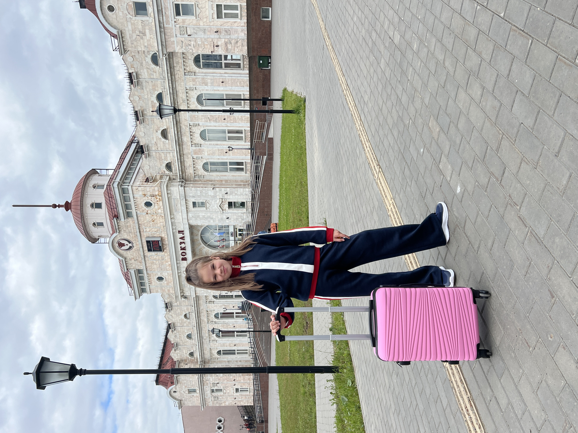 A young girl with blonde hair in a ponytail stands on a paved walkway in front of a building that appears to be a train station, identified by the Russian word "ВОКЗАЛ" (VOKZAL) prominently displayed on its facade. She is dressed in a dark blue tracksuit with red and white accents and is holding the handle of a bright pink wheeled suitcase. The building itself is ornate, with a domed roof and large arched windows, and appears to be constructed of light-colored stone. Several lampposts stand in the foreground and around the building. The sky is overcast. The scene is located in Izhevsk, Russia, likely at the Izhevsk railway station.