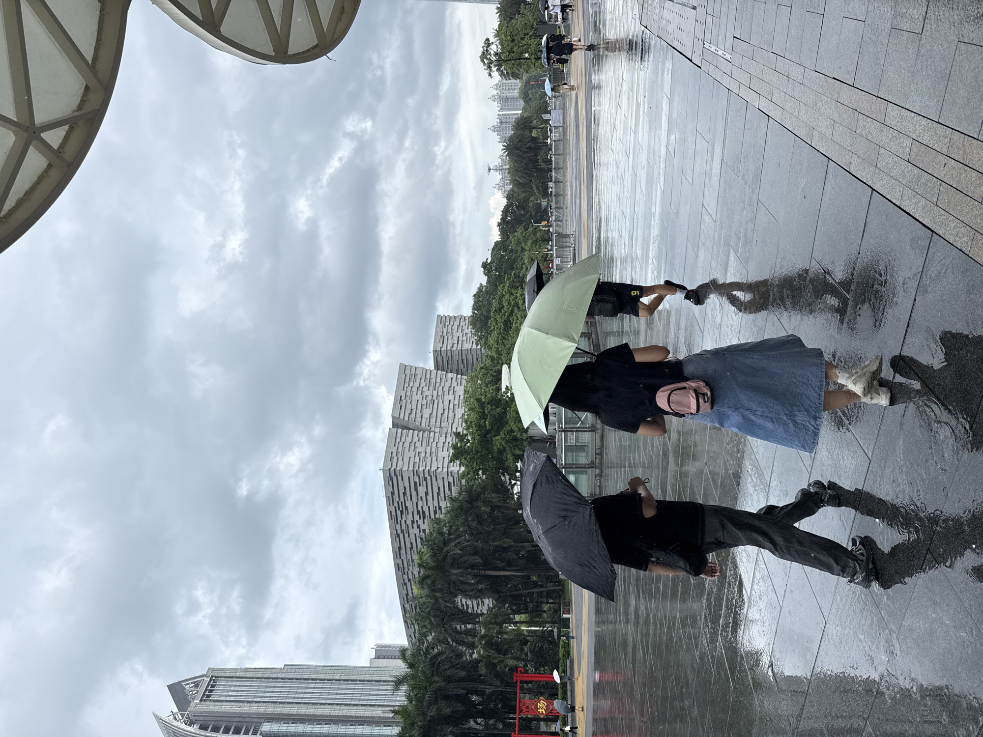 In Tianhe District, China, on a rainy day, three people are walking on a wet, tiled plaza. The person in the foreground to the left is holding a black umbrella and wearing a black t-shirt, dark pants, and athletic shoes. To their right, another person is holding a light green umbrella and wearing a black t-shirt, a light blue denim skirt, and white sneakers. Behind them, a third person is walking with a black umbrella and wearing dark shorts and athletic shoes. The plaza is reflective due to the rain, and palm trees and modern buildings, including a tall skyscraper and a distinctive grey building with a patterned facade, are visible in the background. The sky is overcast with dark clouds.