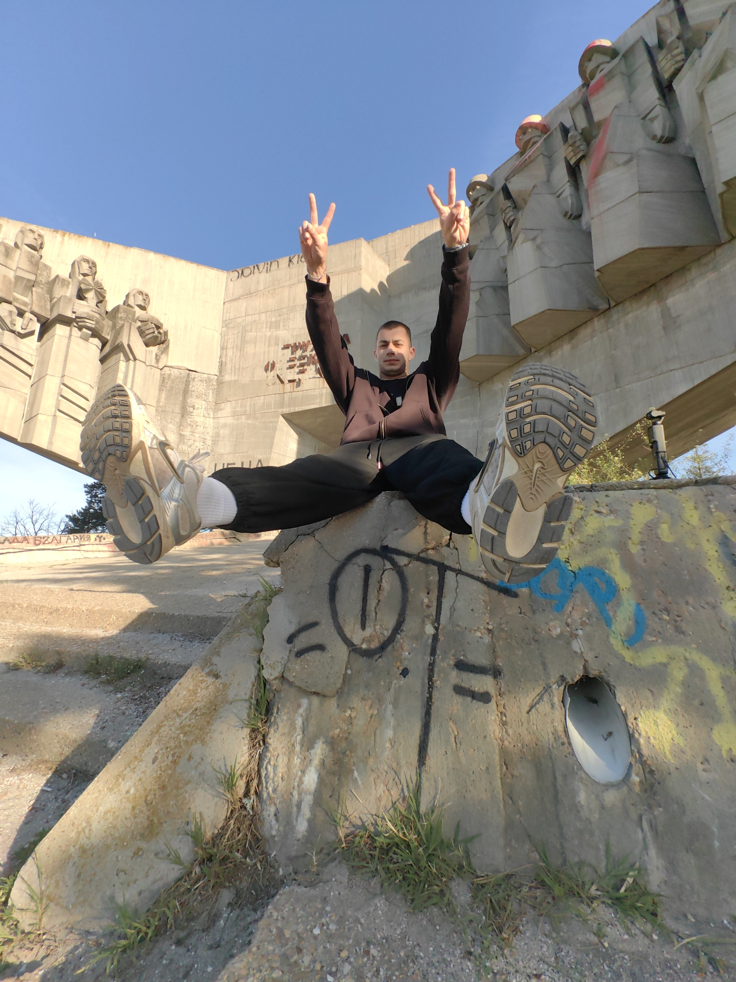 A low-angle full shot depicts a man sitting on a concrete structure with graffiti, with his legs extended upwards, showcasing the soles of his shoes. He is wearing a brown hoodie, black pants, and white socks, and he is holding up both hands in a peace sign gesture.
Behind him stands a monumental concrete structure featuring socialist-era sculptures and Cyrillic lettering. The location is likely the Pantheon of the Renaissance in Varna, Bulgaria. The day is clear and sunny, with a blue sky. Vegetation is visible around the concrete structure.