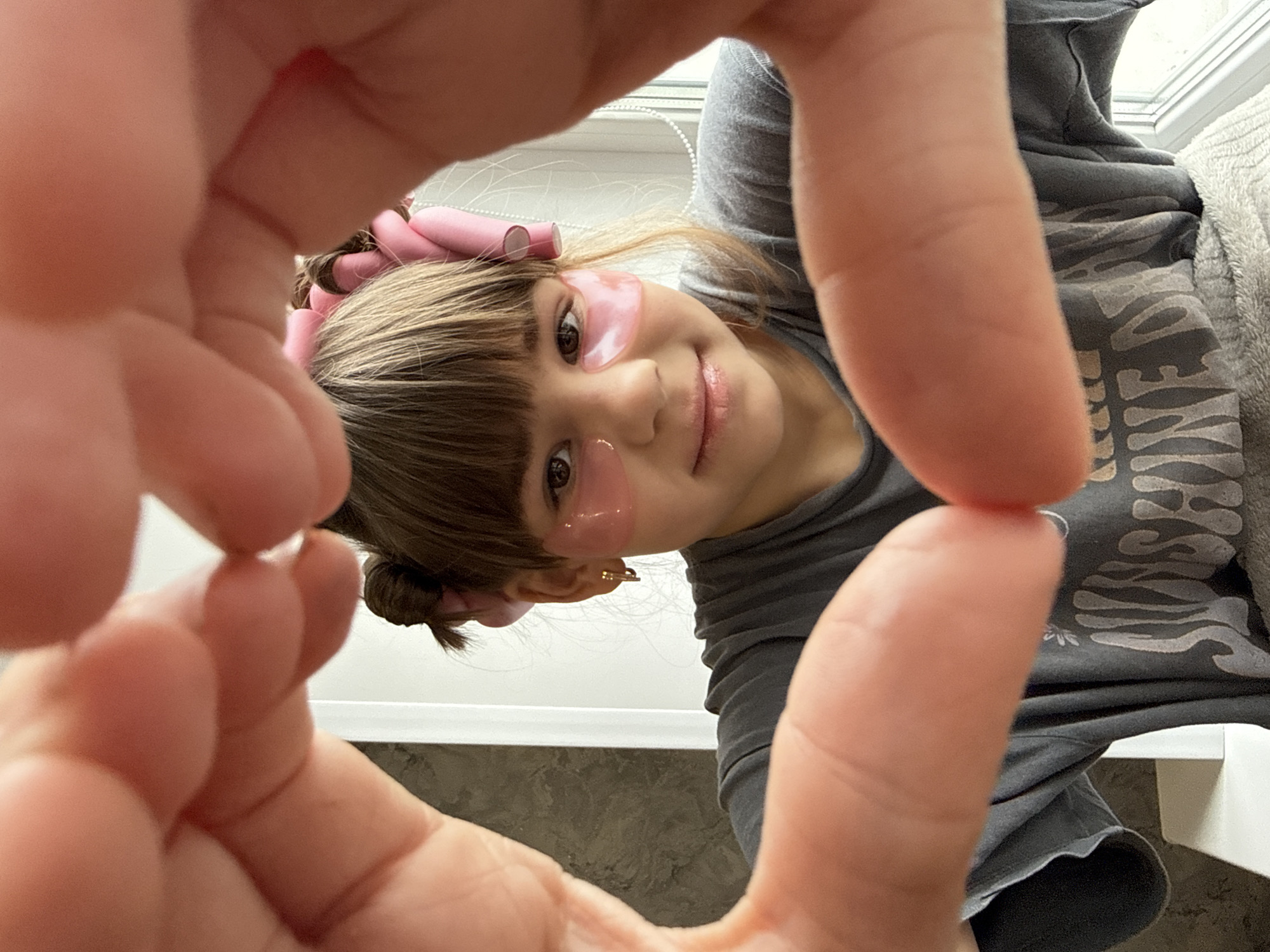 The image presents a close-up, first-person perspective of a young child, whose head is inverted relative to the camera's orientation. The child's face is playfully framed by the out-of-focus hands of the photographer. She has light brown hair with bangs and subtle highlights, with pink hair rollers visible on the sides of her head, indicating a beauty or styling activity. Pink, gel-like under-eye patches are applied beneath both her dark brown eyes. She wears a gentle, closed-mouth smile, conveying a sense of contentment or amusement as she looks directly at the camera.
She is dressed in a dark gray t-shirt with a stylized, light-colored text pattern across the chest that appears to read "WEEKEND VIBES," though parts are obscured. The background is softly lit, likely by a window, suggesting an indoor daytime setting. A light-colored object, possibly furniture or a window frame, is visible behind her. The image captures a candid and endearing moment of self-care or preparation, with the close framing adding intimacy and focus on the child's expression and facial accessories.