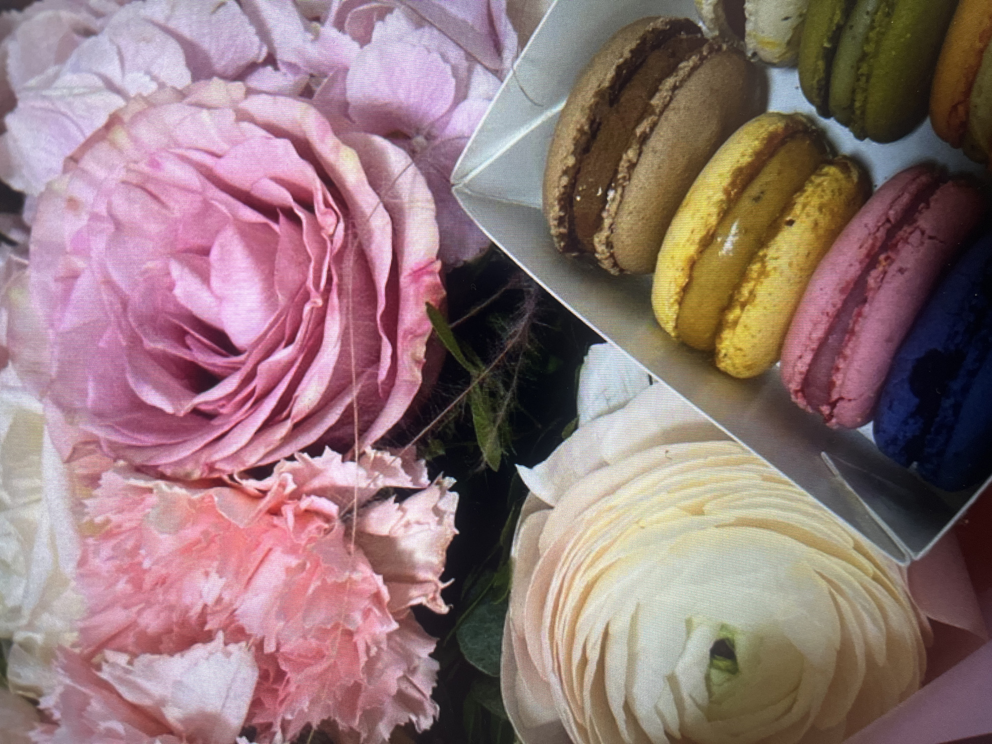 In the foreground of the image, a white cardboard box contains an assortment of colorful macarons. There are at least seven visible macarons: one brown, two yellow, one pink, one dark blue, two green, and one orange. The macarons are arranged in rows within the box.
Behind the box of macarons, a bouquet of flowers is visible. The bouquet features large, delicate pink roses and lighter pink carnations. There is also a pale yellow, layered flower, possibly a ranunculus, in the lower left corner. The flowers are lush and appear to be in full bloom. The background is softly blurred, suggesting a shallow depth of field. The overall lighting is soft and natural.
There is no indication of the location being in Moscow, Russia, or any specific city. The image depicts a still life arrangement of flowers and macarons. There are no people or actions taking place.
