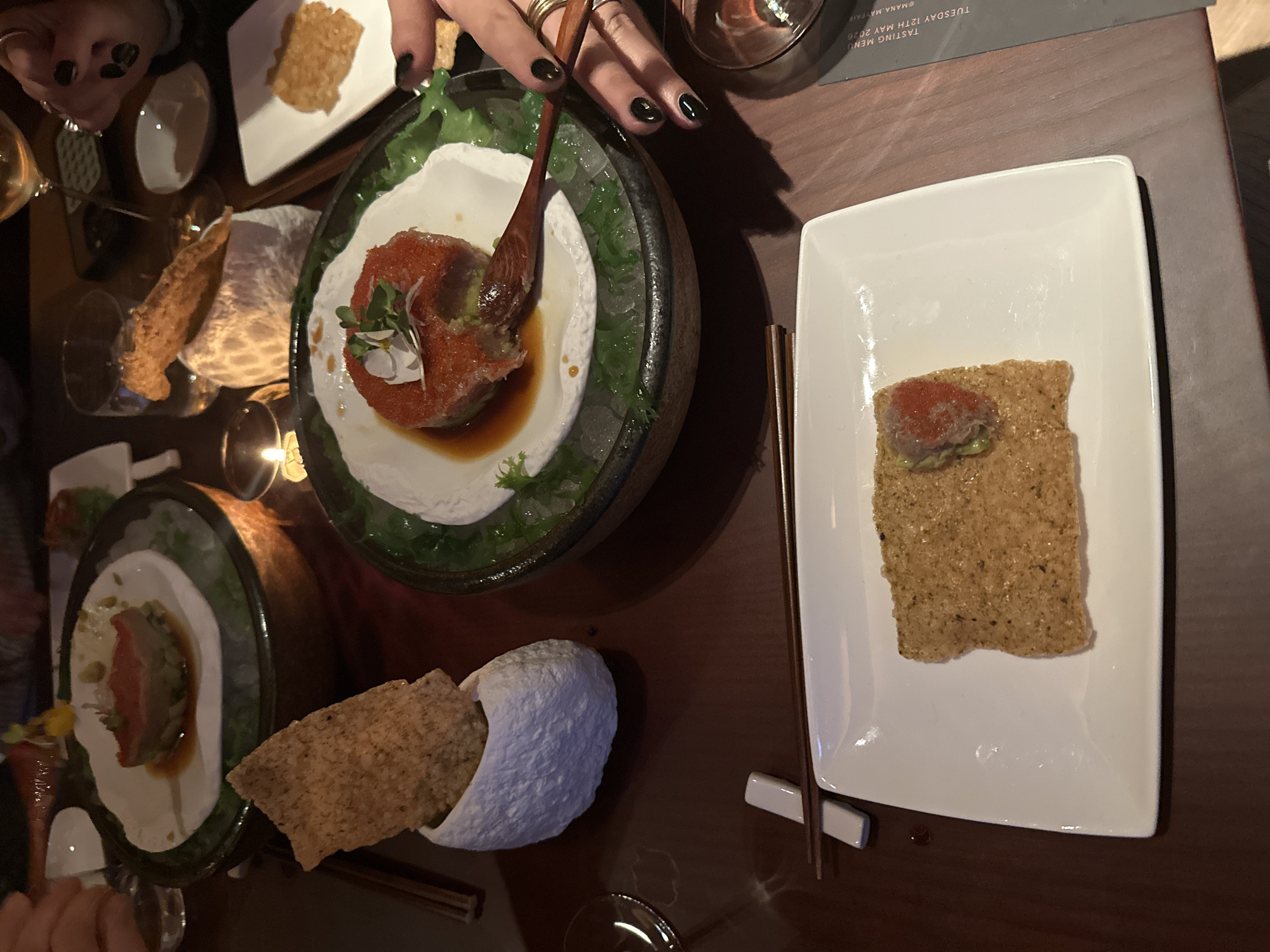 A tabletop scene depicts an array of food dishes. A dark-rimmed bowl contains a white circular base with a red food item garnished with green accents. A white plate to the right features a square cracker topped with similar red and green food. A hand is holding a wooden spoon, presumably interacting with the food. Another dark-rimmed bowl with a similar food presentation is visible, partially obscured. Crackers are displayed in a white cloth or paper container. Chopsticks rest on a white holder next to the white plate. The location is suggested to be the city of London, United Kingdom, evidenced by the tasting menu which reads "@Hana Mayfair, Tuesday 12th May 2026".