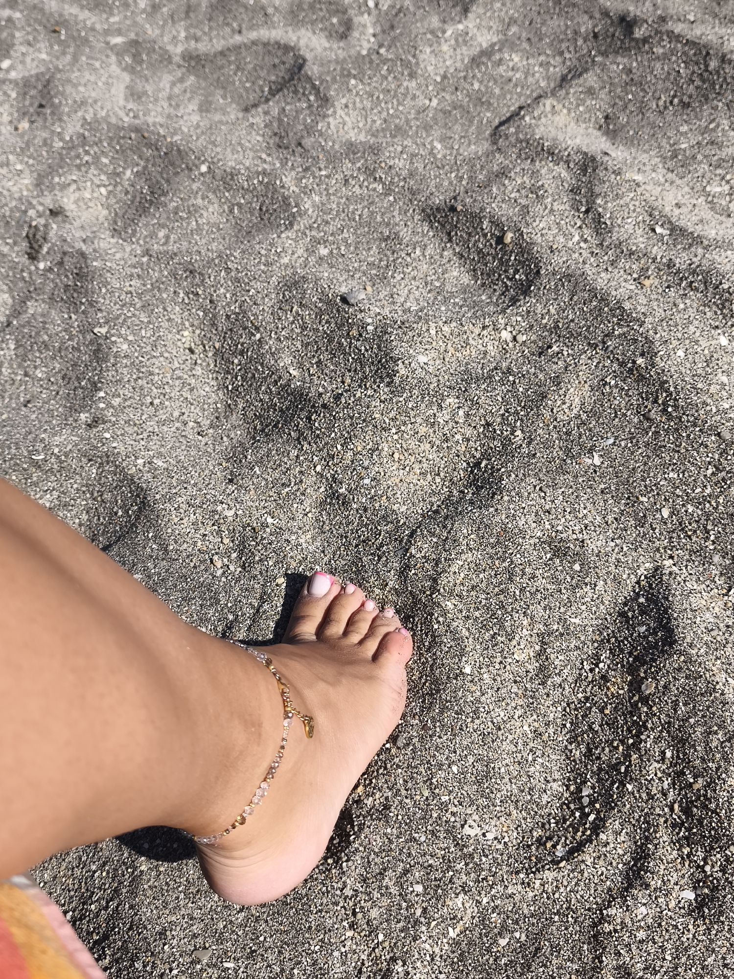 This media file is a close-up, high-angle shot of a person's bare feet and lower leg on a sandy beach. The sand is dark gray and appears coarse, mixed with small white and gray pebbles. Shadows are cast on the sand, suggesting it is daytime, likely sunny. The person's foot is positioned in the lower left quadrant of the frame, with the toes pointing towards the right. The toenails are painted with a light pink and white gradient. A delicate gold anklet with small beads and a charm is visible around the ankle. The skin tone is tanned. The background consists entirely of the sandy beach, with some subtle undulations and textures creating a natural pattern. There is no visible text in the image. The scene evokes a sense of leisure and relaxation, typical of a beach environment. The location context provided indicates this scene is in Burgas, Bulgaria.