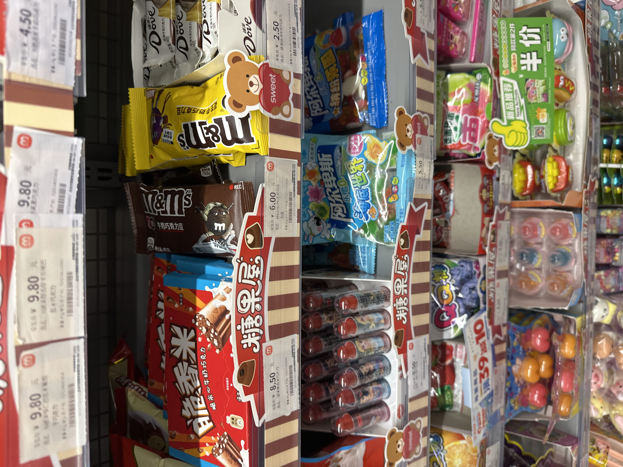 The image depicts a shelf in a store, likely a convenience store or supermarket, in the Chongqing High-tech Zone, China. The shelf is stocked with various candies and chocolates. Prominently displayed are M&M's in yellow and brown packaging, Dove chocolates, and a red box of "脆米心牛奶巧克力" (Crispy Rice Milk Chocolate). Other candies include "阿尔卑斯" (Alps) brand candies in blue packaging. Price tags are visible, displaying prices in yuan. There are bear-shaped decorations around the edges of the candy sections. A promotion for a discount is displayed: "满39减10" (spend 39 get 10 off). The image is brightly lit, with the products neatly arranged on the shelf.