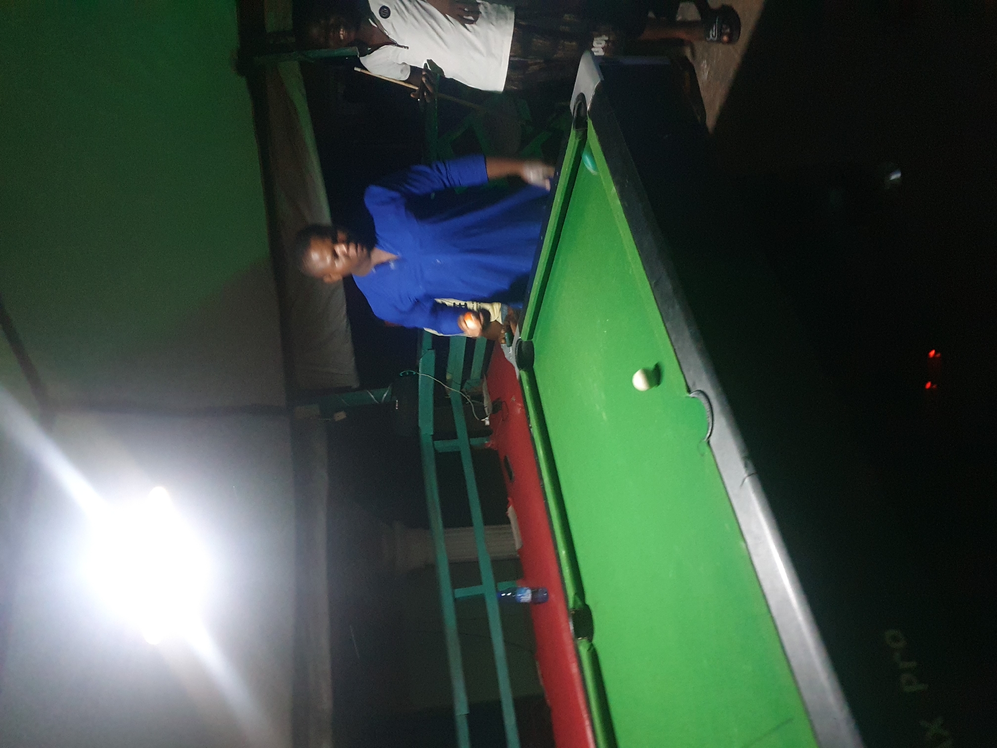 In Lokoja, Nigeria, two men are captured in an outdoor setting, possibly a rooftop or patio, illuminated by a bright overhead light. A pool table dominates the foreground, its green felt surface showing a single white ball. To the left, a bottle of water rests on a red railing. The man on the left, wearing a blue garment, holds an orange ball, appearing to be in the process of playing pool. To the right, another man in a white polo shirt and patterned shorts smiles, holding a cue stick. The background suggests an elevated space with a dark, possibly covered, area above. The overall scene depicts a casual game of pool at night.