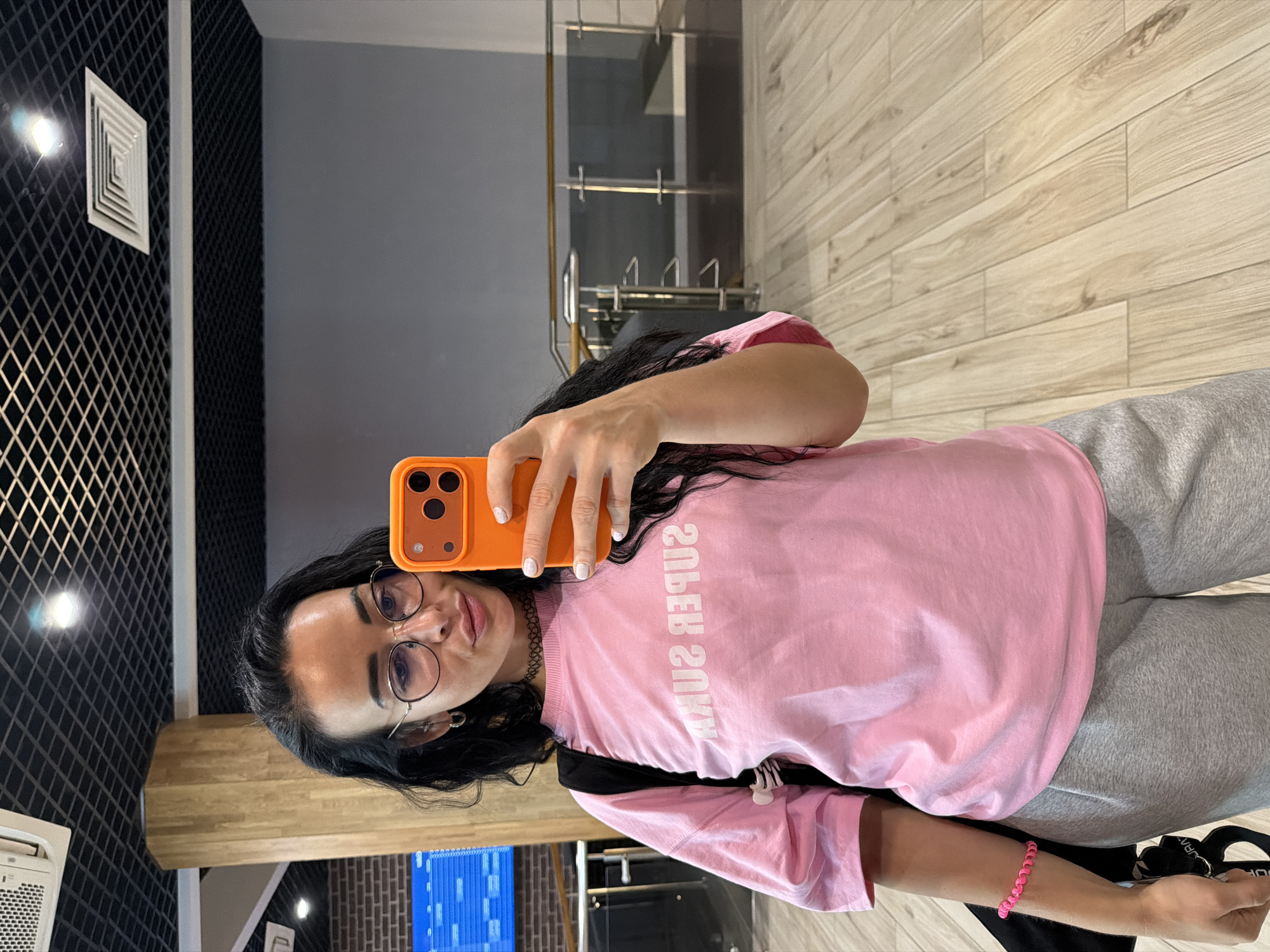 A woman is taking a selfie. She is wearing eyeglasses, a pink shirt with "SUPER SOUA" printed on it, and grey sweatpants. She is holding an orange smartphone in her right hand. Her hair is dark and curly. She is also wearing a pink bracelet on her left wrist.
The background indicates an indoor setting, with elements suggesting a modern or renovated space. There are brickwork and glass elements that suggest a contemporary design. Based on the architectural details in the reflection, this is in Saint Petersburg, Russia.
