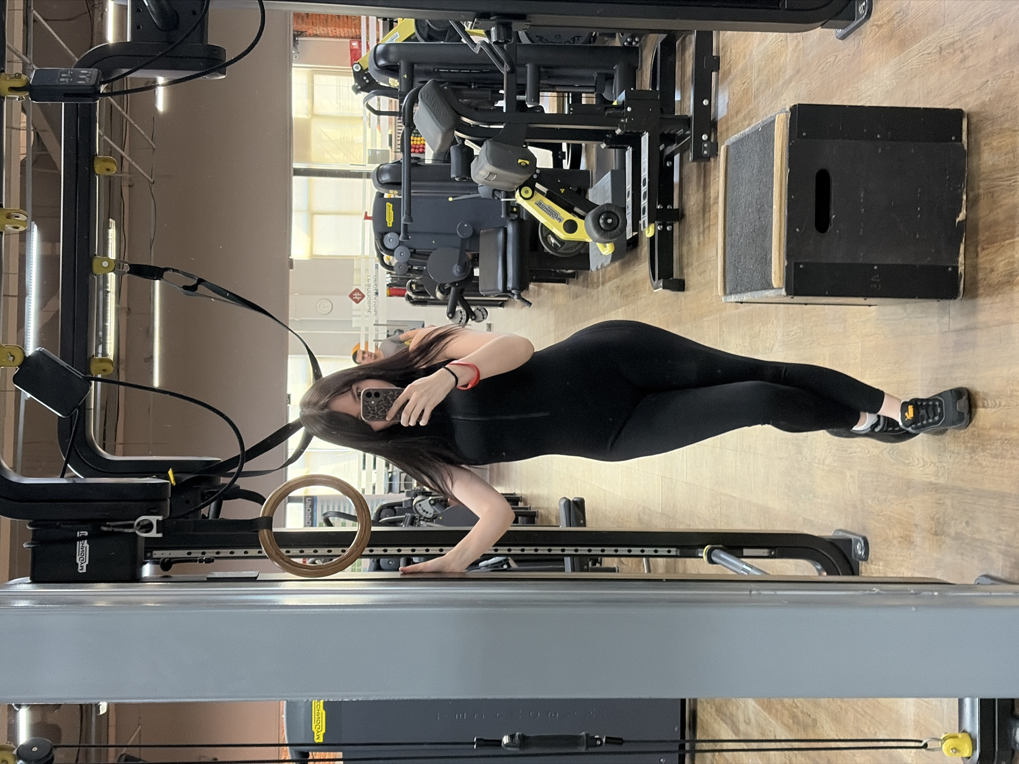 A woman in a black jumpsuit and athletic shoes is taking a selfie in a gym. The gym has weight machines, a wooden box, and gymnastic rings. The location is in the city of Chaikovsky, Russia.