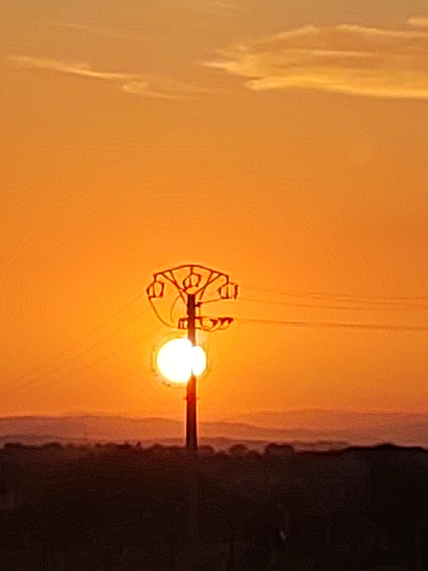 The image depicts a sunset scene with an electric pylon silhouetted against a bright orange sky. The pylon is centrally located and features a distinctive, ornate top structure. A bright white orb, likely the sun, is positioned behind the pylon, partially obscured by its structure and wires. In the foreground, a dark, silhouetted landscape with rolling hills is visible, hinting at the natural environment surrounding Sobradillo, Spain. The sky above the horizon is a gradient of orange and yellow, with faint wispy clouds. The overall impression is one of a serene and dramatic sunset in a rural or semi-rural setting.