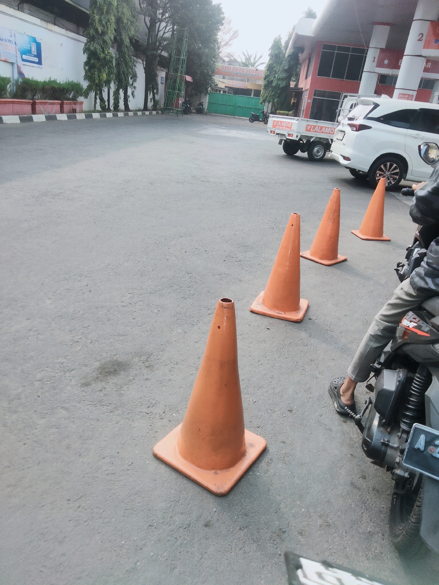 This image captures a street scene in Tangerang, Indonesia, likely near a gas station or an industrial area. In the foreground, four orange traffic cones are strategically placed on an asphalt surface, indicating some form of temporary lane closure or traffic management. To the right, a person is seated on a motorcycle, their legs clad in grey pants and black croc-style footwear. The motorcycle itself is partially visible, with its engine and rear wheel in focus, and a license plate starting with "A 2..." is discernible.
In the mid-ground, a white pickup truck with the "Lalamove" logo is parked, facing left. Next to it, a white SUV is also parked. Beyond these vehicles, a red and white building, possibly a gas station with visible pump numbers "1" and "2," stands prominently. Several trees line the area, and a green metallic structure, resembling scaffolding or a tower, is positioned near the building. In the background, a white wall with a blue advertisement is visible, along with other buildings and greenery, contributing to a typical urban Indonesian landscape. The overall lighting suggests it is daytime, with some overcast conditions.