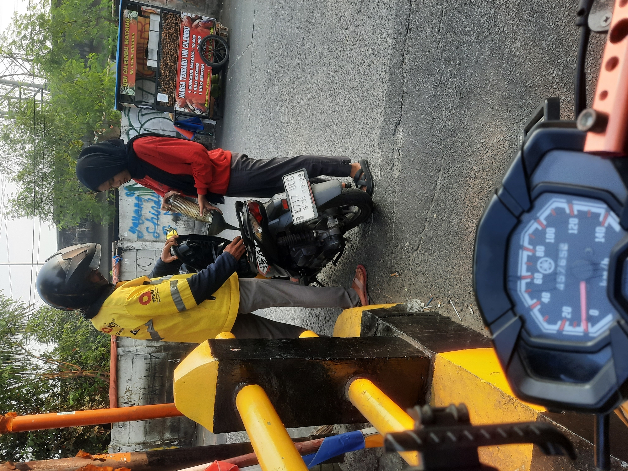 A street scene in North Jakarta, Indonesia, features a male delivery person wearing a yellow jacket with "YPSSI" and "Gojek" branding, and a black helmet. He is standing next to a motorcycle and interacting with a woman wearing a black hijab and a red long-sleeved shirt. The woman is pouring liquid from a metal cylinder into a funnel held by the delivery person, who is placing it into the motorcycle's fuel tank. In the background, a food cart selling "Ubi Cilembu Matang" is visible, with prices listed for different quantities of sweet potatoes. The environment includes a concrete wall with graffiti, trees, and a paved road with a speed bump. The perspective is from the rider of another motorcycle, capturing the dashboard in the foreground.