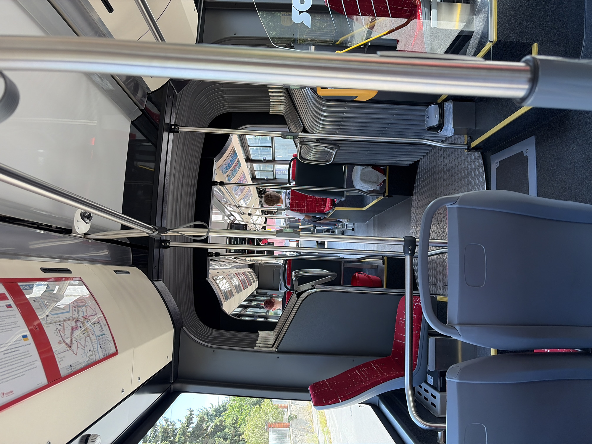 The image depicts the interior of a modern bus, likely a public transit vehicle, with a focus on the seating area and aisle. The color palette is predominantly gray and red, with metallic accents from handrails and structural elements.
In the foreground, two gray seats are visible, facing forward. Beyond these, the aisle leads towards the rear of the bus. To the left, a red seat with a patterned design is prominent, positioned near a window that offers a view of trees and a fence, suggesting an urban or suburban environment outside.
Further down the aisle, several passengers are seated. One person, seen from the back with reddish-brown hair, is sitting in a red seat. Another passenger, also viewed from behind, is wearing a white shirt and is seated in a red seat further back. The seats appear to be made of a durable fabric, with metal framing.
Various functional elements of the bus are visible, including vertical metal poles for passengers to hold onto, luggage racks above the windows, and what appear to be route maps or advertisements displayed on the ceiling. A red bucket is situated near the aisle, and a small trash can with a lid is also present. The flooring is a dark material, with a metal grate pattern in some areas.
The lighting appears to be a mix of natural light from the windows and internal bus lighting, creating a well-lit interior. The overall impression is one of a clean and orderly public transportation vehicle. While the image itself does not explicitly state its location, given the context of a previous query specifying Nitra, Slovakia, it is reasonable to assume this scene is within the city of Nitra, on a bus route serving the local population.