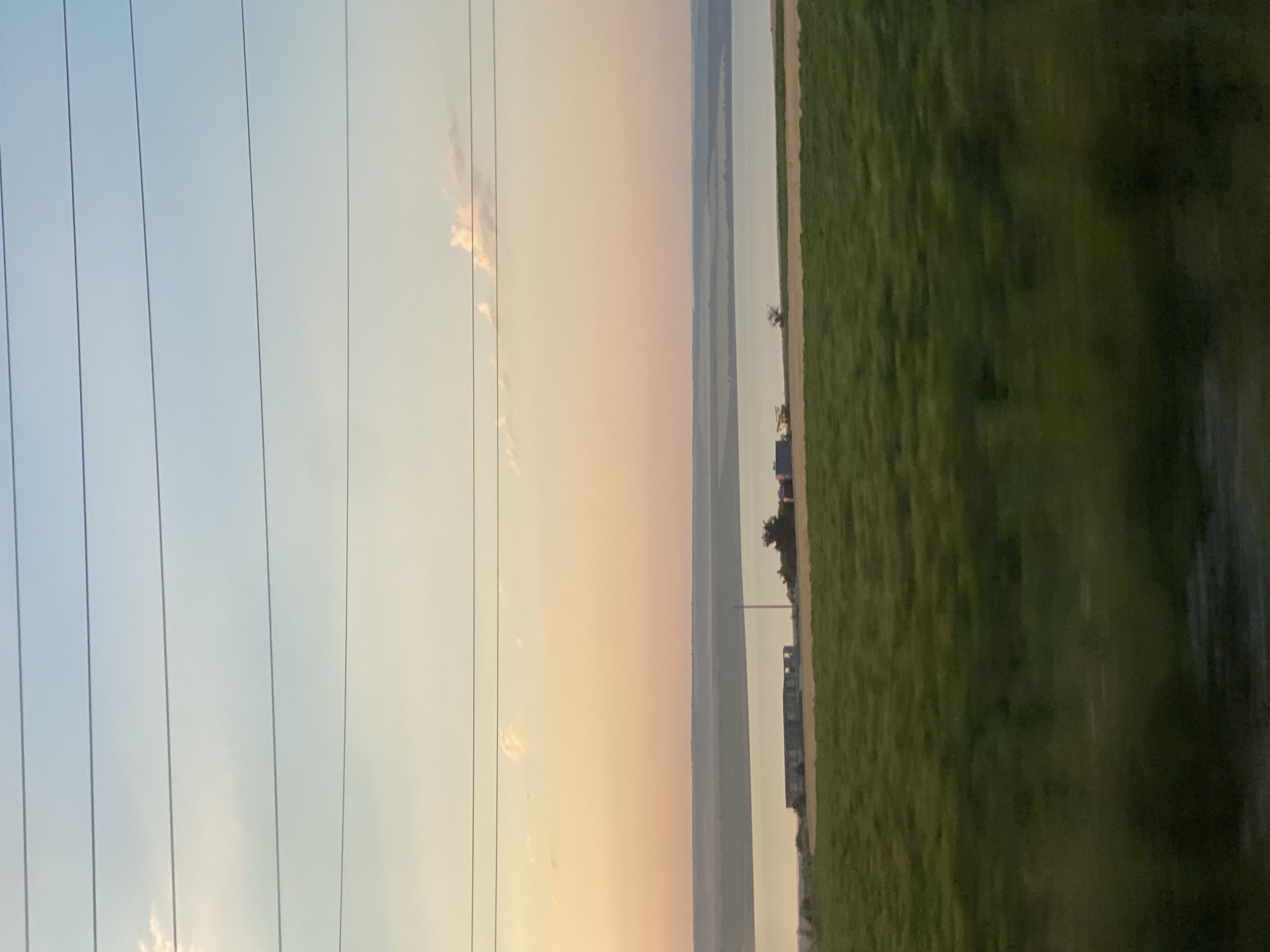The image depicts a rural landscape at sunset, featuring a vast body of water and cultivated fields. The sky transitions from a pale blue at the top to hues of orange and pink near the horizon, with a few scattered clouds. Several parallel power lines stretch across the upper portion of the frame. In the middle ground, a body of water, likely a river or lake, separates the foreground from a distant shore. Along the edge of the water, several structures are visible, including what appear to be modular buildings or shipping containers, along with some vegetation and a solitary pole. The foreground is dominated by a large, grassy field, appearing slightly blurred due to motion, suggesting the photo was taken from a moving vehicle. The scene is devoid of human presence. The overall impression is one of tranquility and natural beauty at the end of the day.