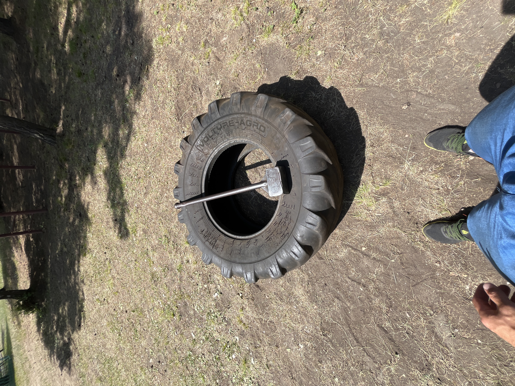 An outdoor scene in Krasnoturansk, Russia, shows a large tractor tire lying flat on the ground. A sledgehammer is placed inside the tire, with its head resting on the inner rim. The ground surrounding the tire is covered in dry grass, dirt, and scattered pinecones. In the foreground, the lower legs and feet of a person wearing blue jeans and athletic shoes are visible, suggesting the perspective of someone standing over the tire. The background features trees and dappled sunlight filtering through their leaves, creating shadows on the ground.
