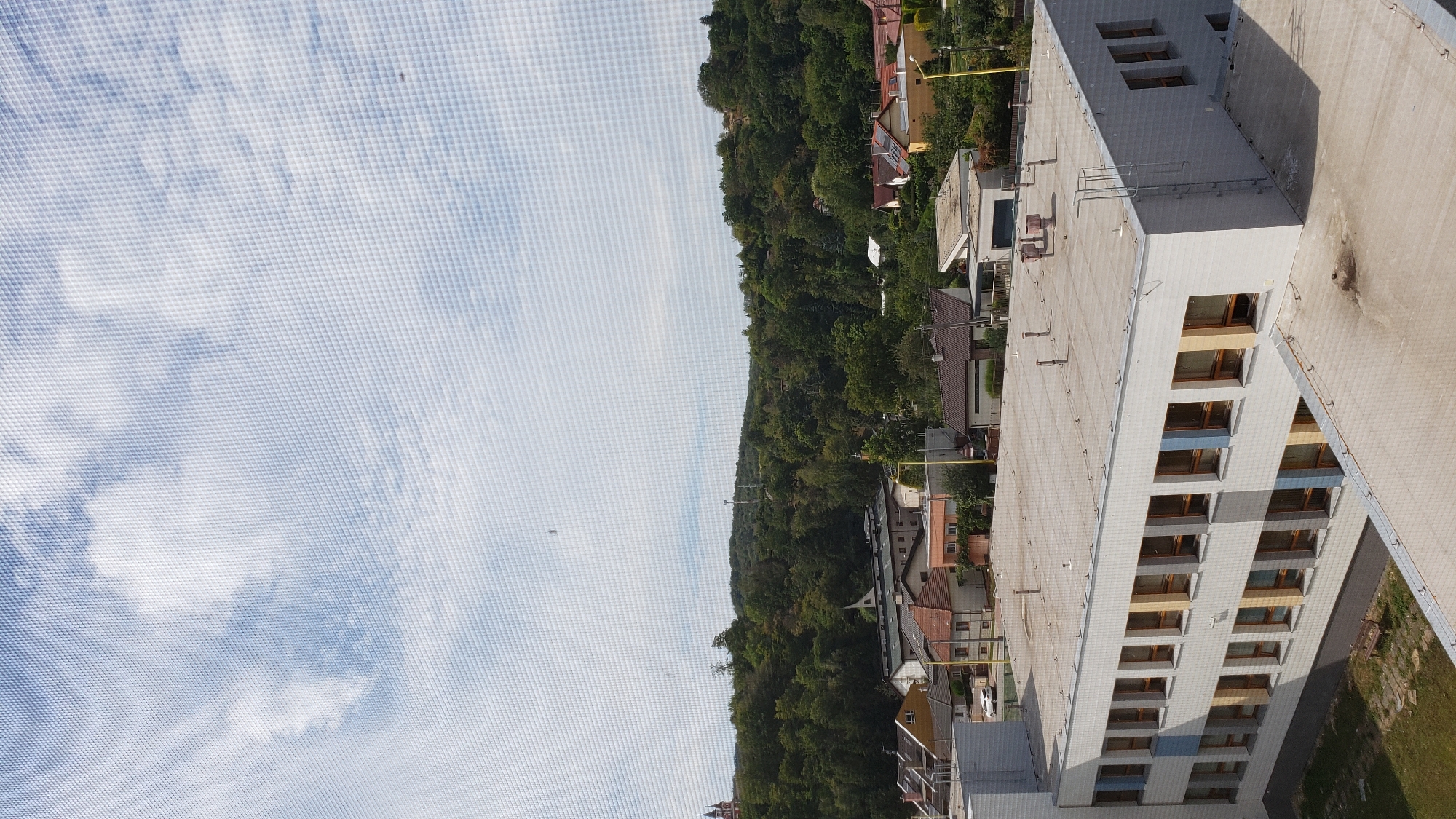 The image shows a view from a high window looking out over a residential area and a wooded hillside in Prešov, Slovakia. In the foreground, the rooftop of a modern, multi-story building with a facade of white tiles and colored window frames is visible. The building has a flat roof with ventilation pipes and other rooftop equipment. Beyond this building, a cluster of houses with red-tiled roofs are visible, interspersed with trees. Further in the background, a dense, green forest covers rolling hills. A few communication towers can be seen on the hills. The sky is partly cloudy with a light blue hue. The overall scene depicts a suburban or semi-rural environment.