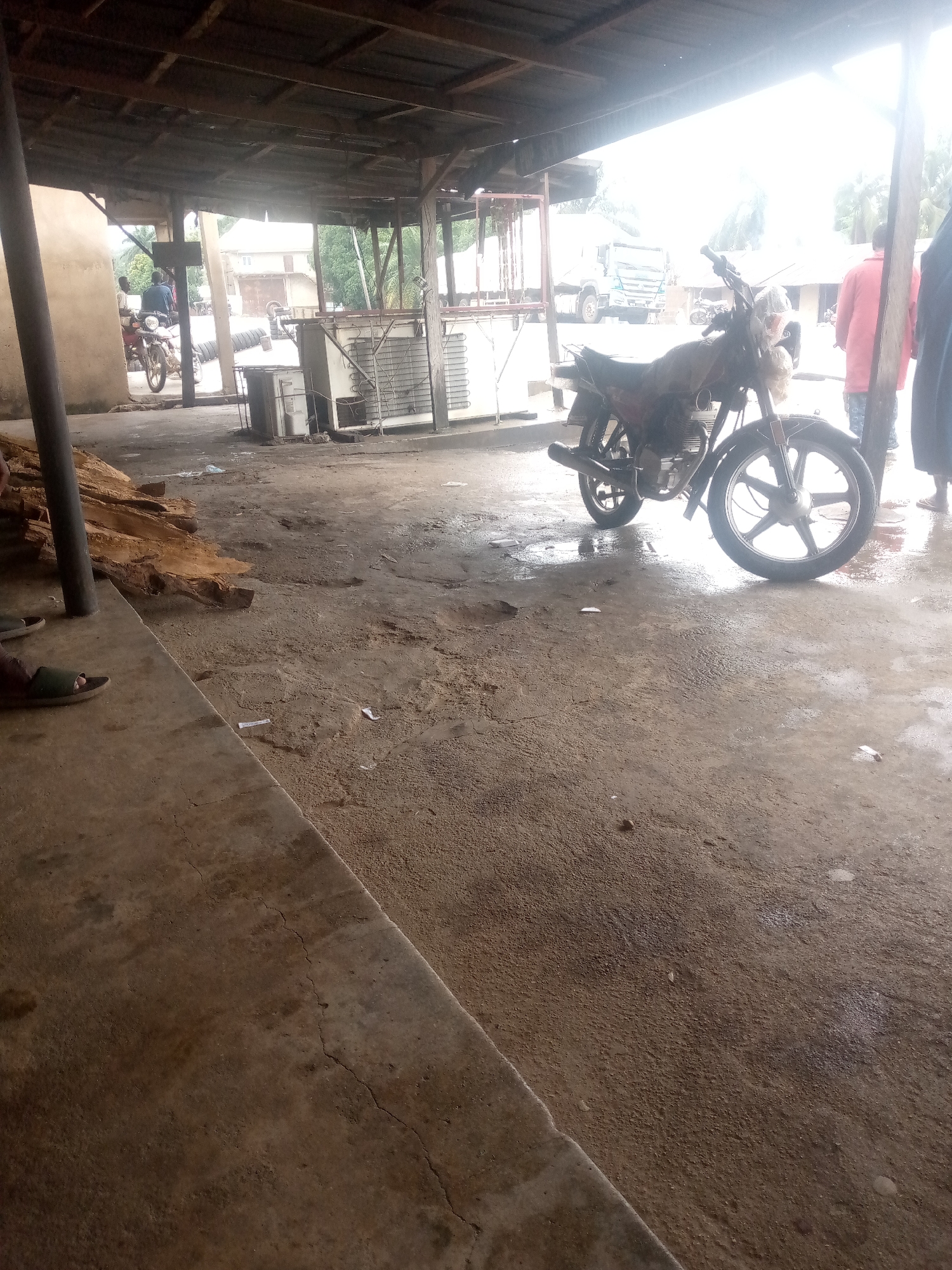 A motorcycle is parked on a wet, concrete floor under a covered, open-air structure. The structure has a wooden roof supported by poles. In the background, a truck is visible, along with several buildings and trees. To the left of the motorcycle, a pile of large, rough-cut wooden logs is present. A person wearing a red shirt and blue shorts stands to the right of the motorcycle. The scene appears to be an outdoor market or workshop in Serti, Nigeria, with evidence of recent rainfall.
