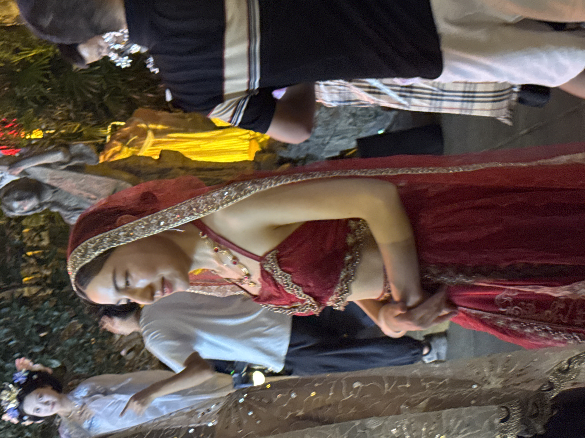 In the Yuzhong District of China, an individual in traditional Indian attire, a red and gold embellished costume with a matching veil, is the central focus of the image. They are positioned in the foreground, facing to the right and slightly away from the camera, with their hands clasped at their waist. Behind them, another person in a light blue garment and a floral headdress is visible, looking to the left and gesturing. Further in the background, several other individuals are present, including one wearing a black and white striped shirt and white shorts, and another in a white t-shirt and dark pants. The scene appears to be outdoors, possibly at an event or festival, given the presence of decorative elements like statues and illuminated foliage, suggesting a festive or cultural setting.
