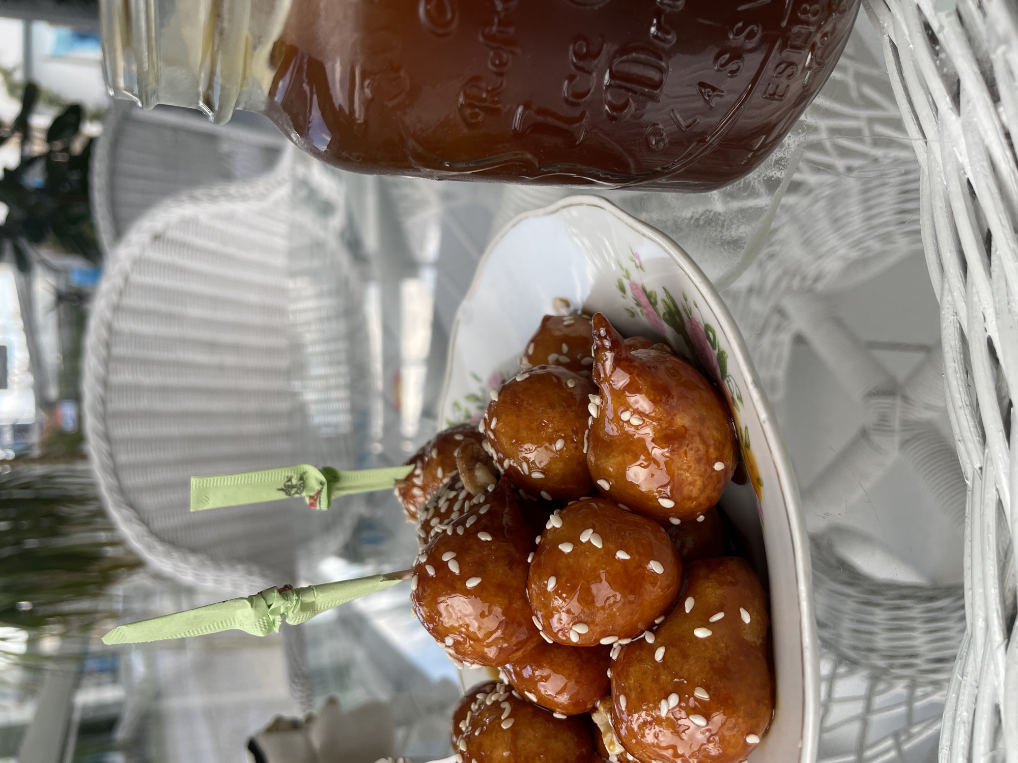 The image shows a plate of loukoumades, Greek doughnuts, glazed with syrup and sprinkled with sesame seeds, with two of the doughnuts speared on decorative green sticks. Beside the plate is a jar of what appears to be honey or syrup. The scene is set on a white wicker table, likely outdoors, as suggested by the natural light and the blurry, out-of-focus background with hints of foliage and buildings. There are no people visible in the image. Although the location is requested as Podgorica, Montenegro, there is no direct evidence in the image to confirm that precise location.