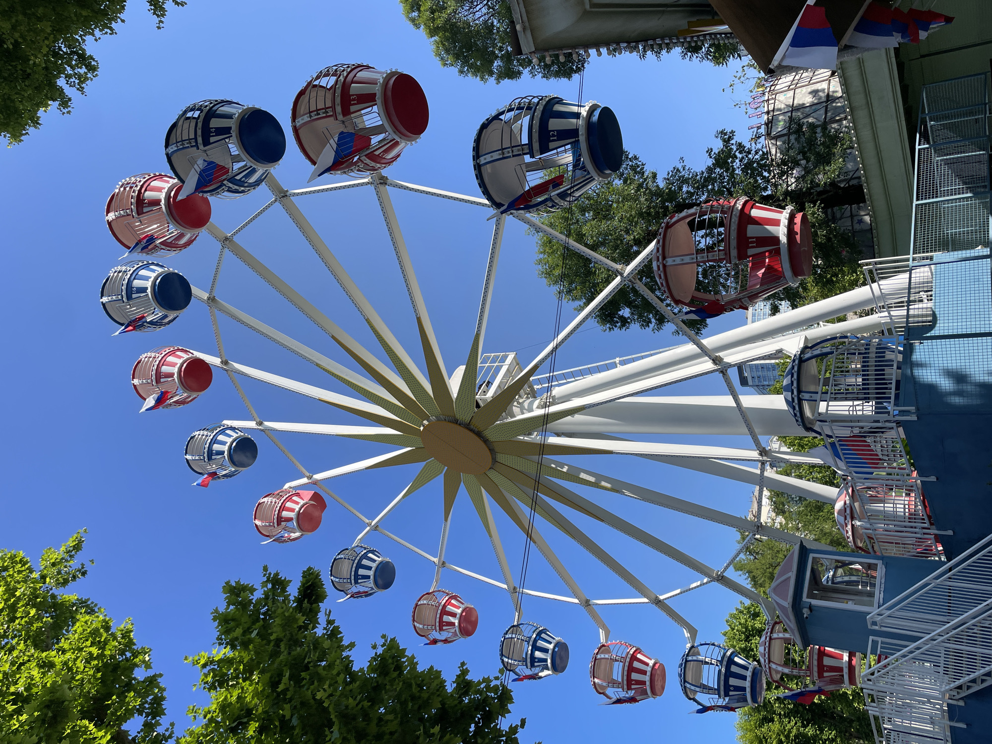 The image shows a Ferris wheel in an amusement park, located in the city of Sochi, Russia. The Ferris wheel is white with a yellow, star-shaped centerpiece. It has multiple gondolas, alternating in red and blue with white stripes. Each gondola is decorated with small Russian flags. The Ferris wheel is set against a clear blue sky, with lush green trees visible in the foreground and background. In the lower portion of the image, a blue and white loading platform for the Ferris wheel can be seen, with stairs leading up to it. The overall scene depicts a sunny day at an amusement park, likely during a season with good weather. There are no people visible in the image.