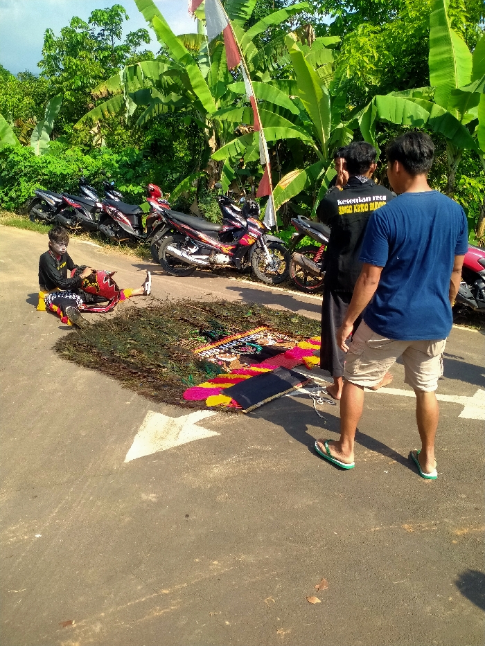 The scene is set on a paved road adjacent to a densely vegetated area in Gempolan, Indonesia. On the left, one individual, with face paint, is seated on the asphalt, dressed in black and white patterned attire with red and black costume elements. This person holds a red and black object, possibly a prop, and wears white sneakers. Five motorcycles are parked off the road behind this individual, partially obscured by thick green foliage, including banana plants. In the center of the road, a pile of green vegetation is arranged next to several colorful, patterned mats or fabric pieces, featuring intricate designs in red, yellow, black, and blue. To the right, two men stand facing away from the camera. The man in the foreground wears a blue t-shirt, light shorts, and green flip-flops. The man behind him wears a black long-sleeved shirt with white text ("KESENIAN REOG..." visible) and dark pants. Both appear to be observing the items on the road and the seated individual. A red and white flag, characteristic of Indonesia, is visible on a pole amidst the foliage. A white directional arrow is painted on the asphalt road surface, pointing left. The area is well-lit by sunlight, casting shadows.