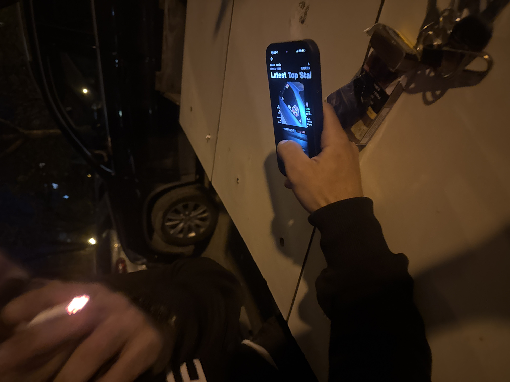 A person holds a lit cigarette in their left hand and a smartphone in their right hand. The phone displays a webpage titled "Latest Top Stal" with an image of a blue car. The person is standing next to a white vehicle with chrome trim, which is likely a car. The background shows a dark street at night. The location is Пловдив, Bulgaria.