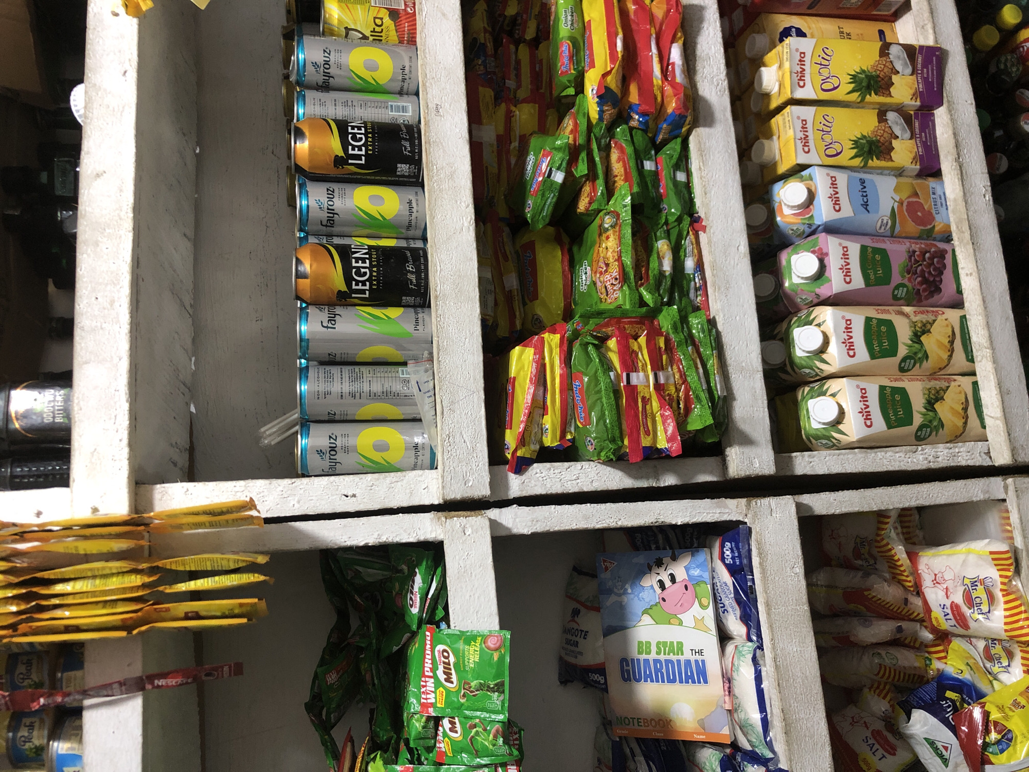 The image shows a shelf in a small shop, likely a convenience store or a corner shop, in Ado, Nigeria. The shelves are stocked with various food items and beverages. On the top shelf, there are cans of Fayrouz pineapple flavor, Legend Extra Stout, and other canned drinks. Some yellow sachets are hanging from the top shelf. Below that, there are packets of noodles, likely Indomie, and other packaged snacks. On the second shelf from the bottom, there are bags of sugar, a children's book titled "BB Star the Guardian," and other packaged goods. The bottom shelf displays more packaged food items, including sachets of salt and what appear to be spices or seasoning. On the right side of the bottom shelf, there are cartons of Chivita juices, including Pineapple Juice, Red Grape Juice, Citrus Mix, and Exotic Pineapple & Coconut flavor. The shop appears to be well-stocked, with items neatly arranged on the shelves. The lighting is somewhat dim, suggesting it might be indoors or late in the day.